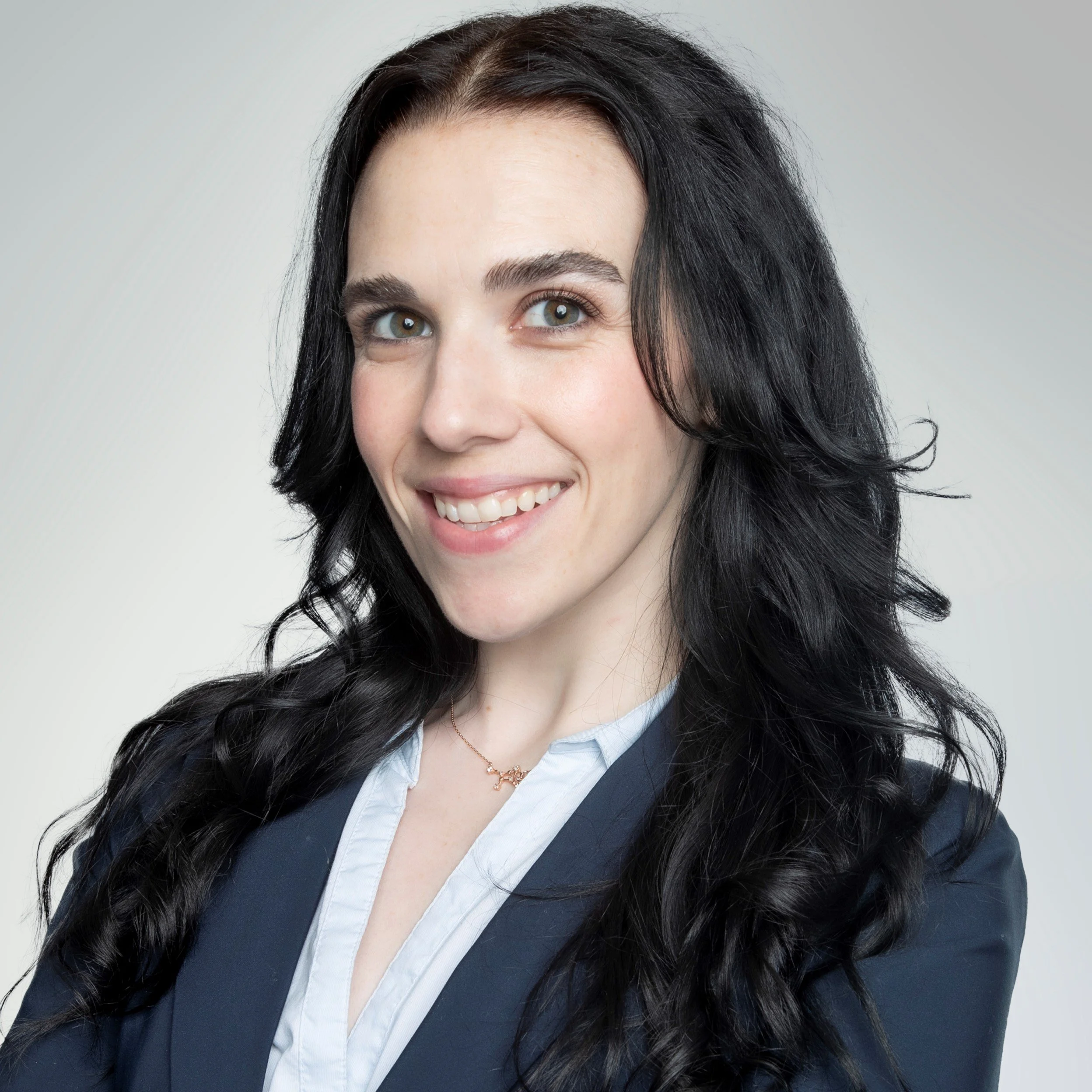 Professional woman with long black wavy hair smiling, wearing a navy blazer and white shirt, against a plain light background.