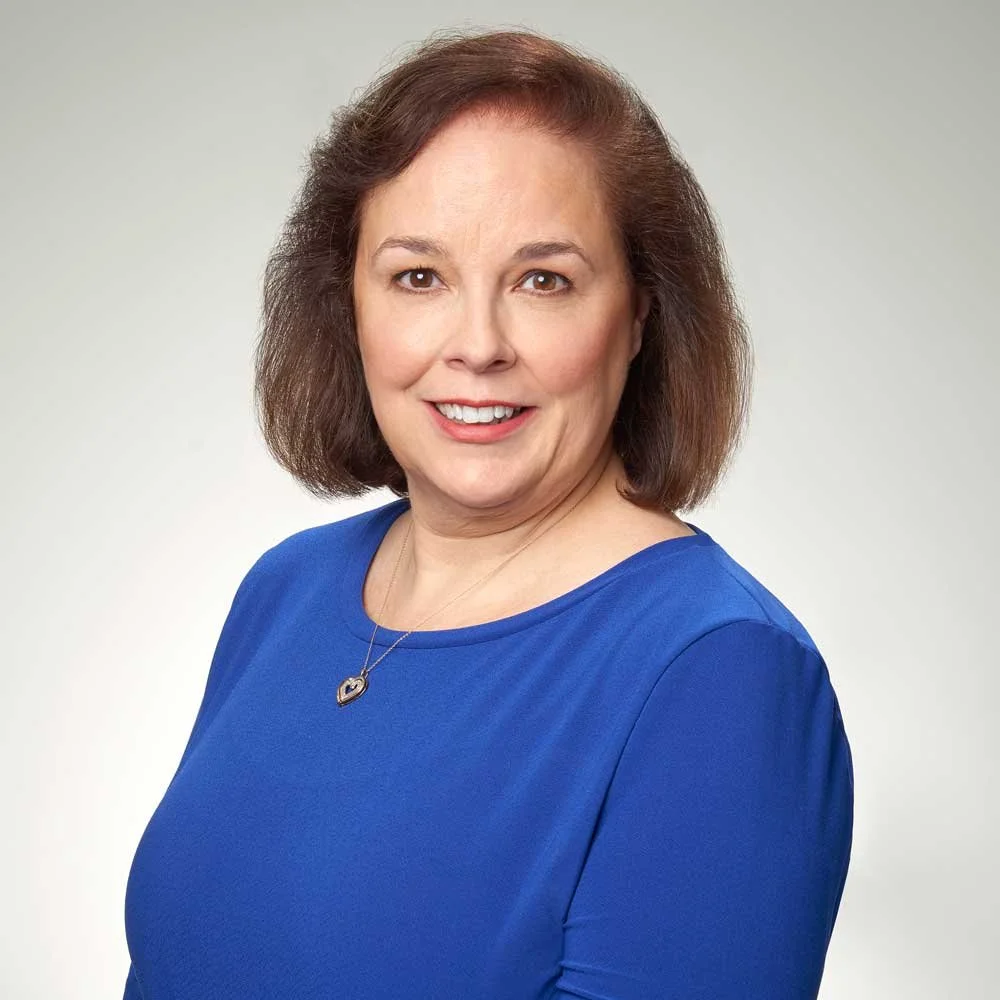 A woman with shoulder-length brown hair, wearing a blue top and a heart-shaped necklace, smiling at the camera against a plain light background.