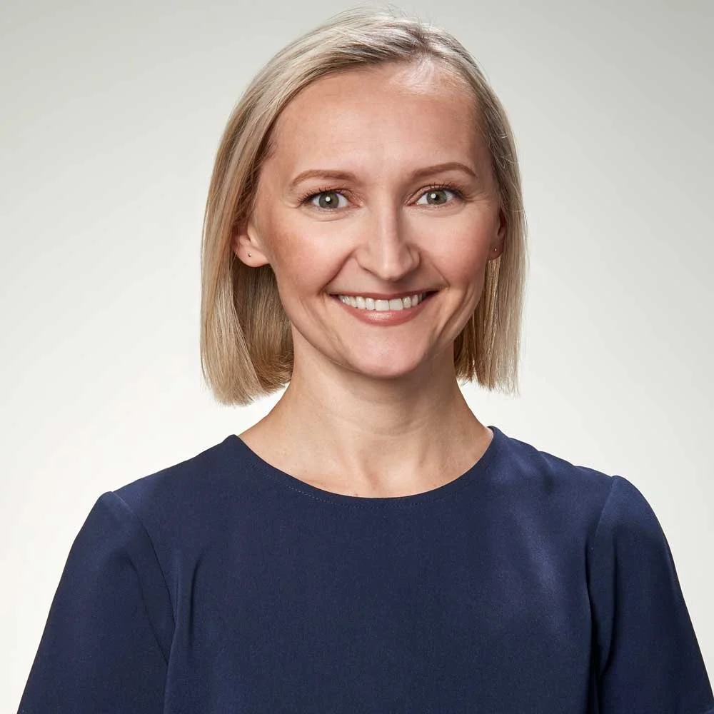 A woman with shoulder-length blonde hair, wearing a navy blue top, smiling against a plain light background.