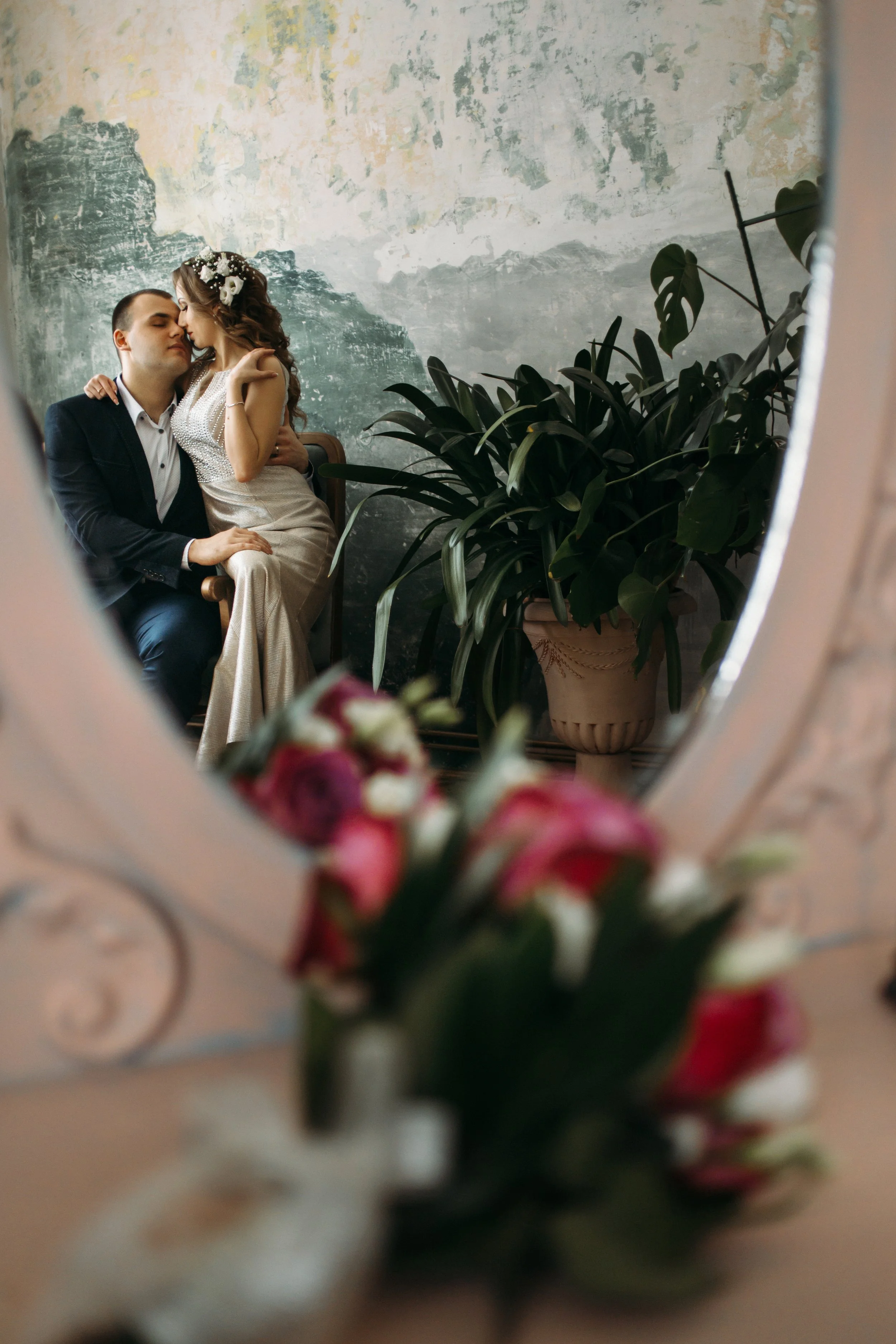 Couple sitting close together, reflected in a mirror, with a blurred bouquet of flowers in the foreground.