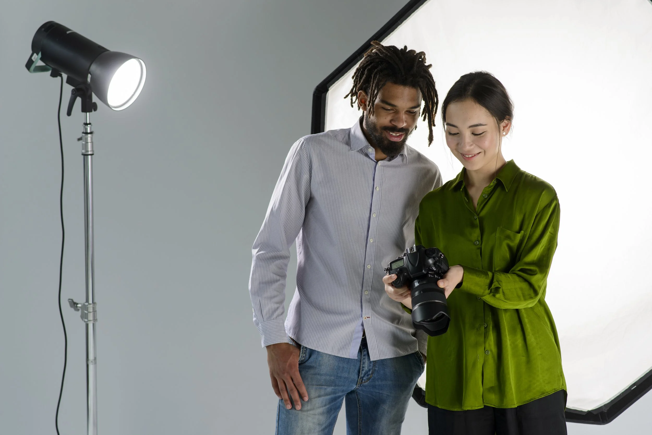 Two people in a photography studio looking at a camera. One man with dreadlocks and a light-colored shirt, and a woman in a green satin blouse.