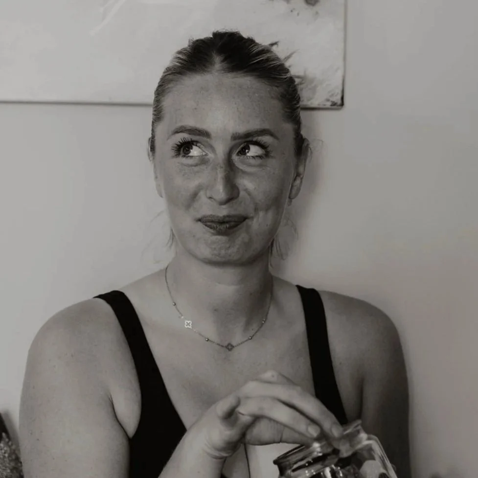 A woman with light hair tied back, wearing a black tank top and a delicate necklace, holding a jar with her right hand, making a playful facial expression with her eyes looking up and to the side.