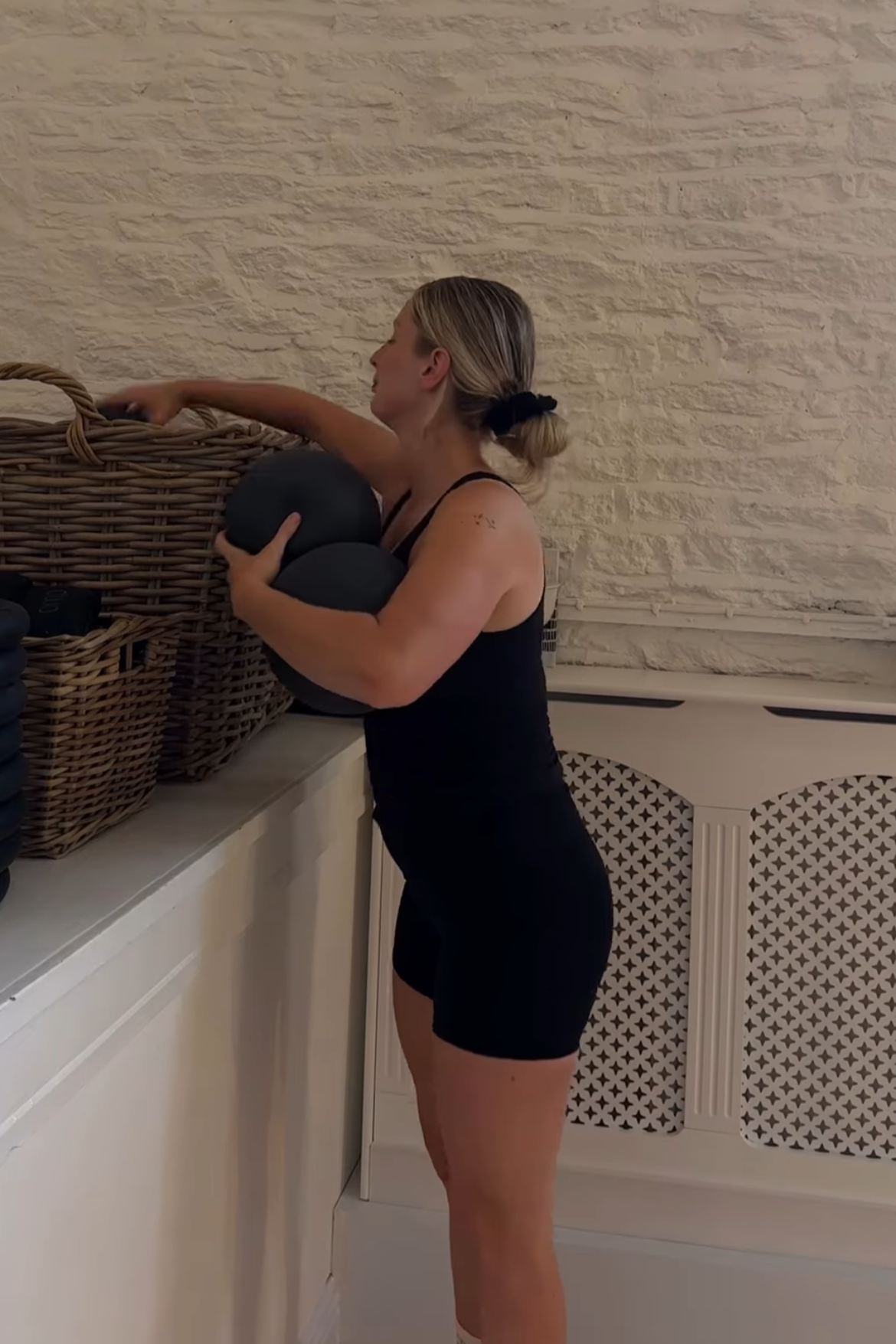 A woman in workout attire reaching into a basket of fitness equipment, holding black exercise balls, in a room with a textured white brick wall and a decorative white radiator cover.