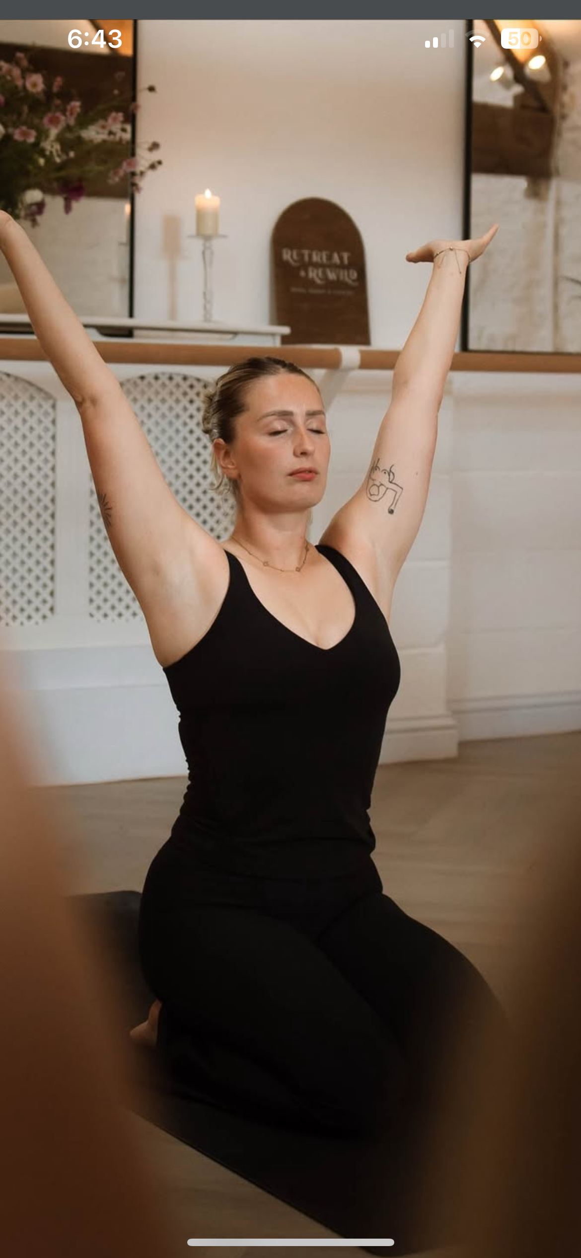 A woman in black athletic clothing practicing yoga with arms raised, kneeling on a mat in a room with a white brick wall and decorative signs and candles in the background.