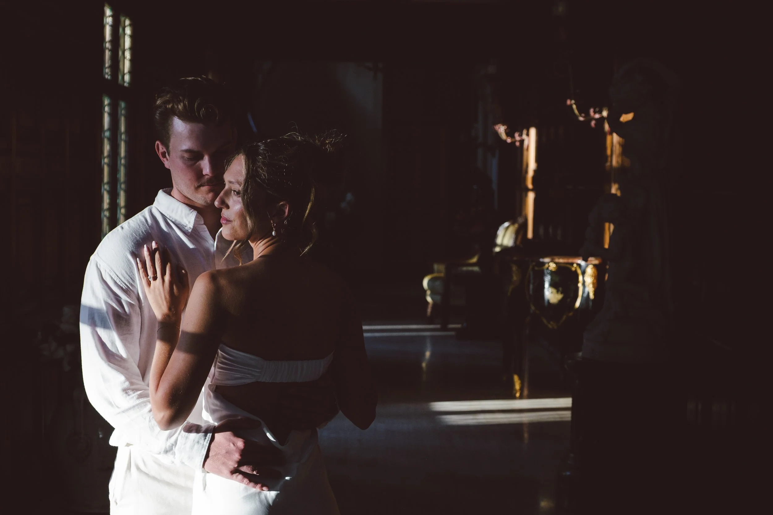 Natural light pours through tall windows as they share a quiet, close moment by the curtains.