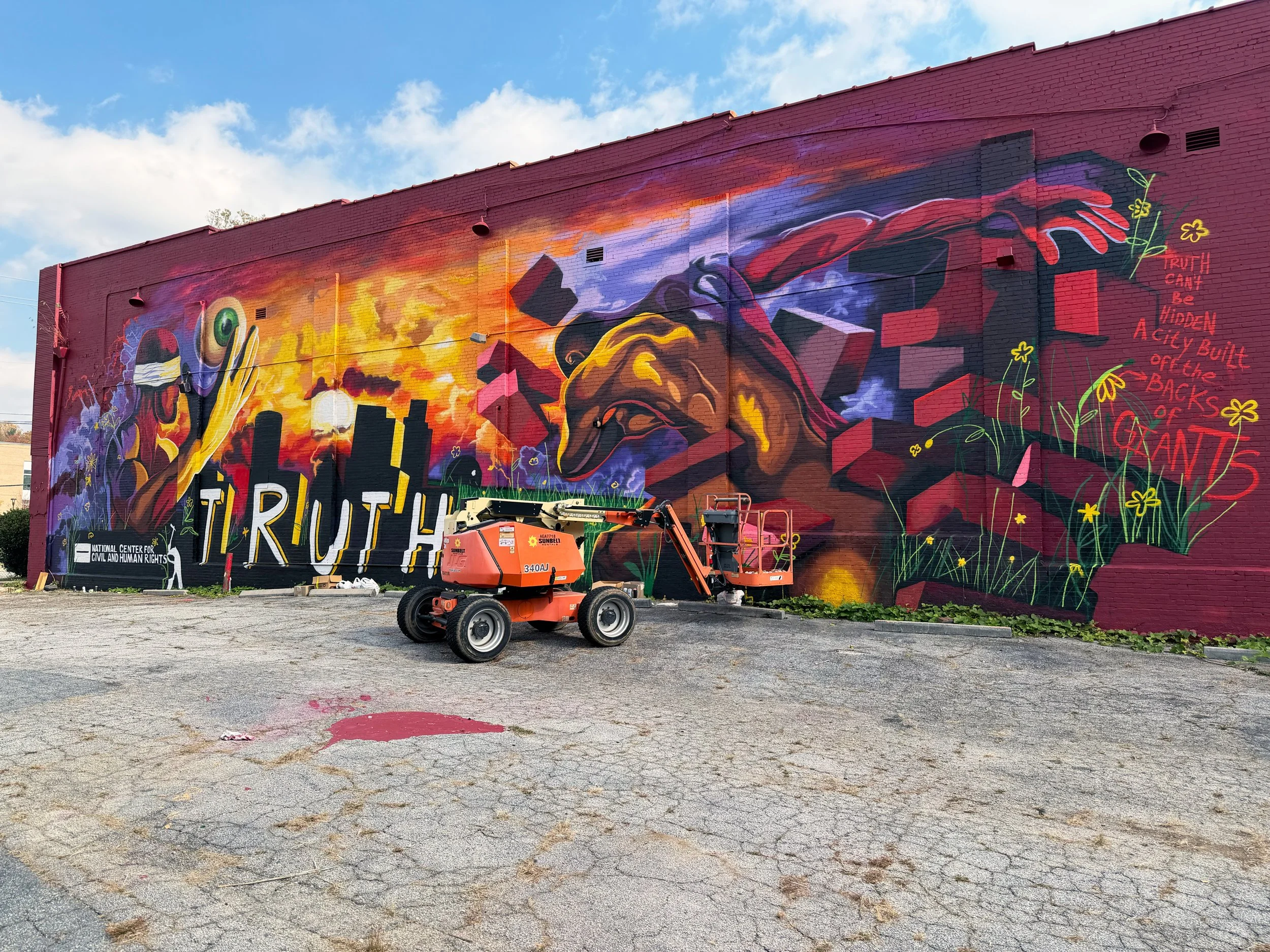 Colorful mural on a brick wall featuring a large gorilla head, a city skyline, vibrant clouds, and flowers. The mural includes the words 'TRUTH' and a quote about truth and secrecy, with a cherry picker vehicle in front of it.