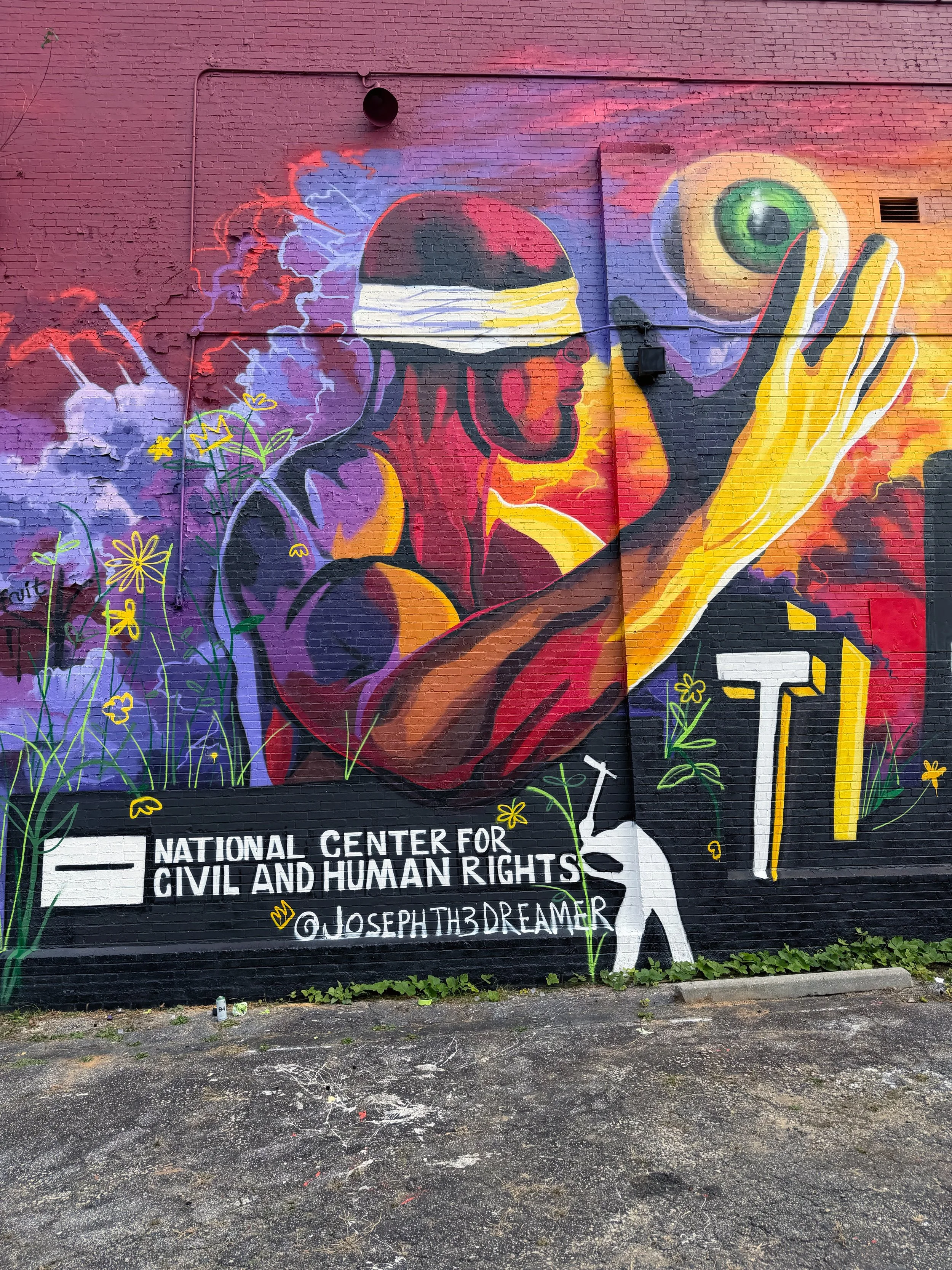 Colorful mural of a person with dreadlocks and a blindfold, holding a large eye, surrounded by flowers and lightning, with text at the bottom reading 'National Center for Civil and Human Rights' and an Instagram handle '@josephth3dreamer'.
