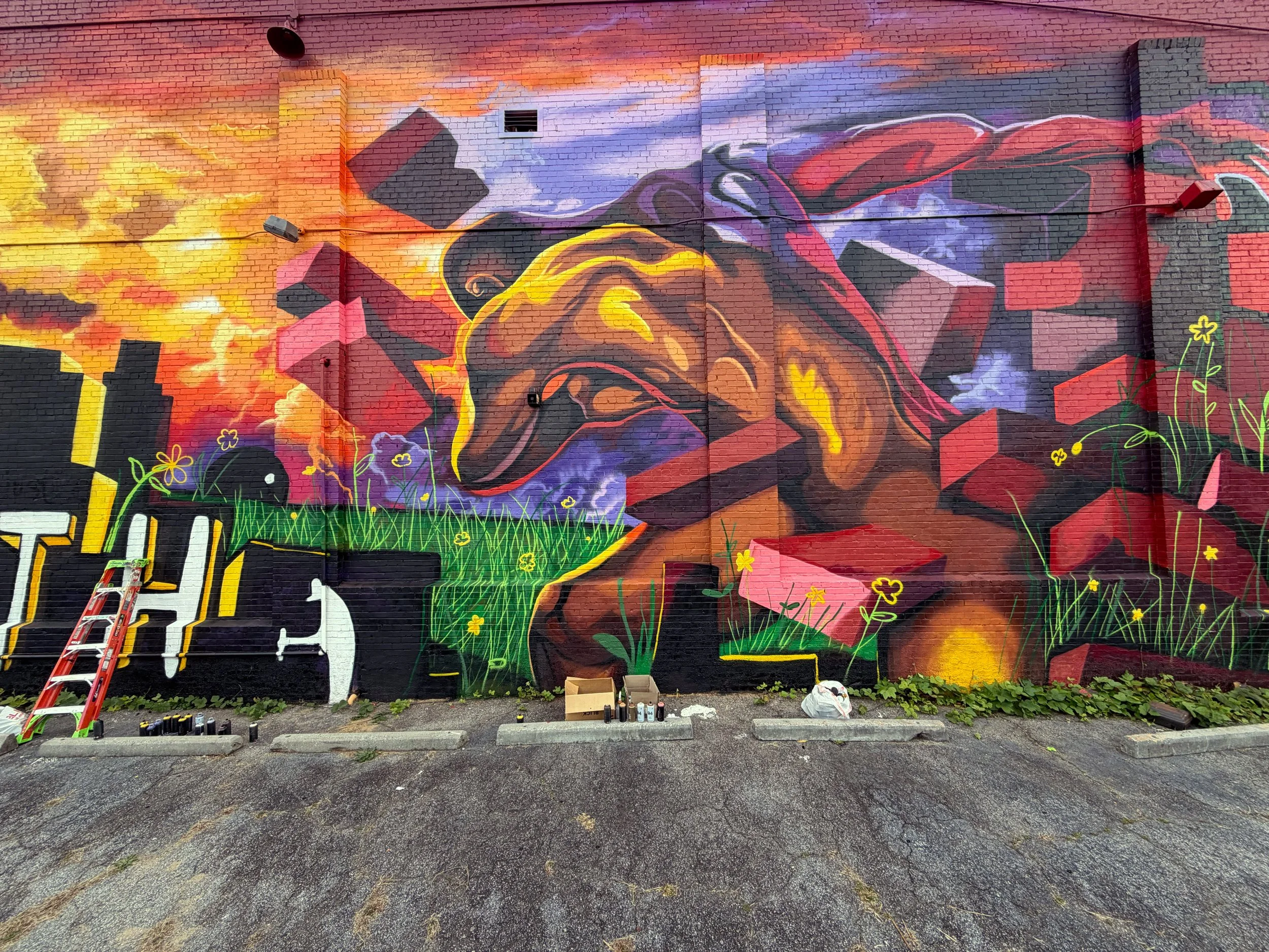 Colorful mural of a dog with a city skyline and sunset in the background, featuring grass, flowers, and red brick elements.