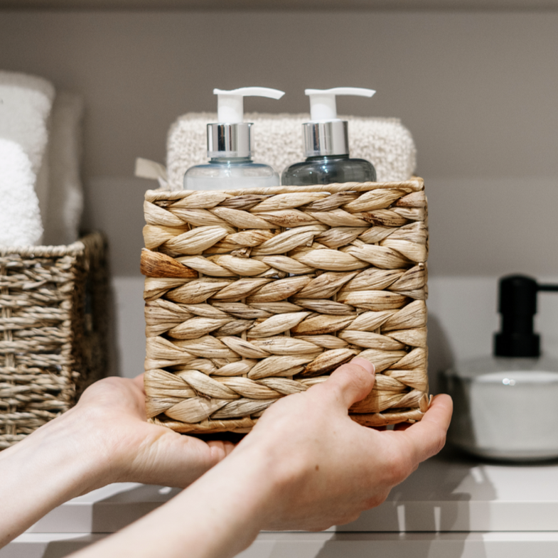 Hands holding a woven basket with toiletries on a bathroom shelf.