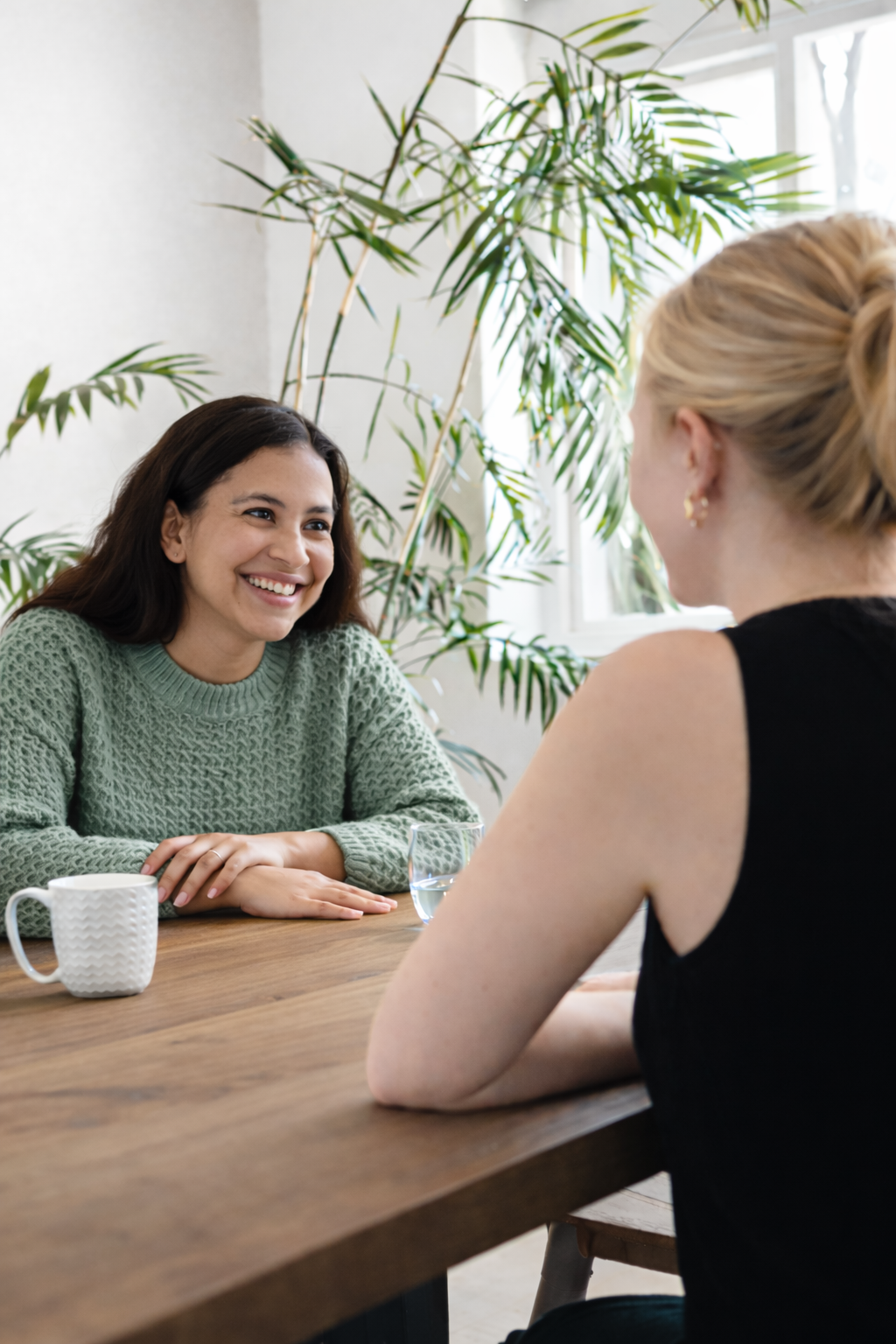 Two women sitting at a wooden table having a conversation in a bright room with large windows and green plants, smiling.