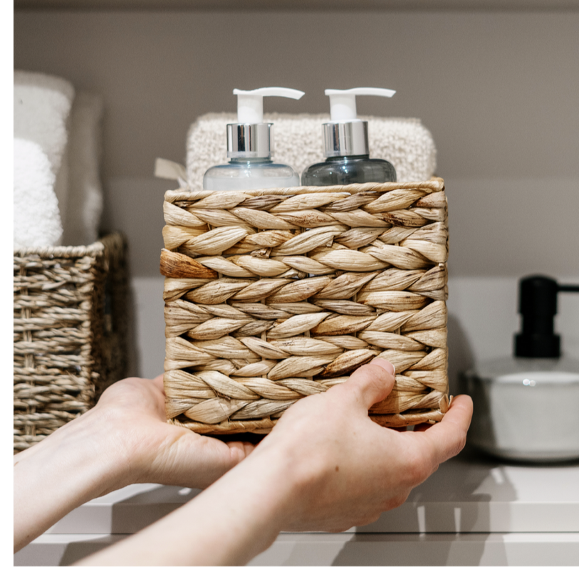 Person placing a woven basket with two soap dispensers on a bathroom counter, with towels and a soap dish nearby.