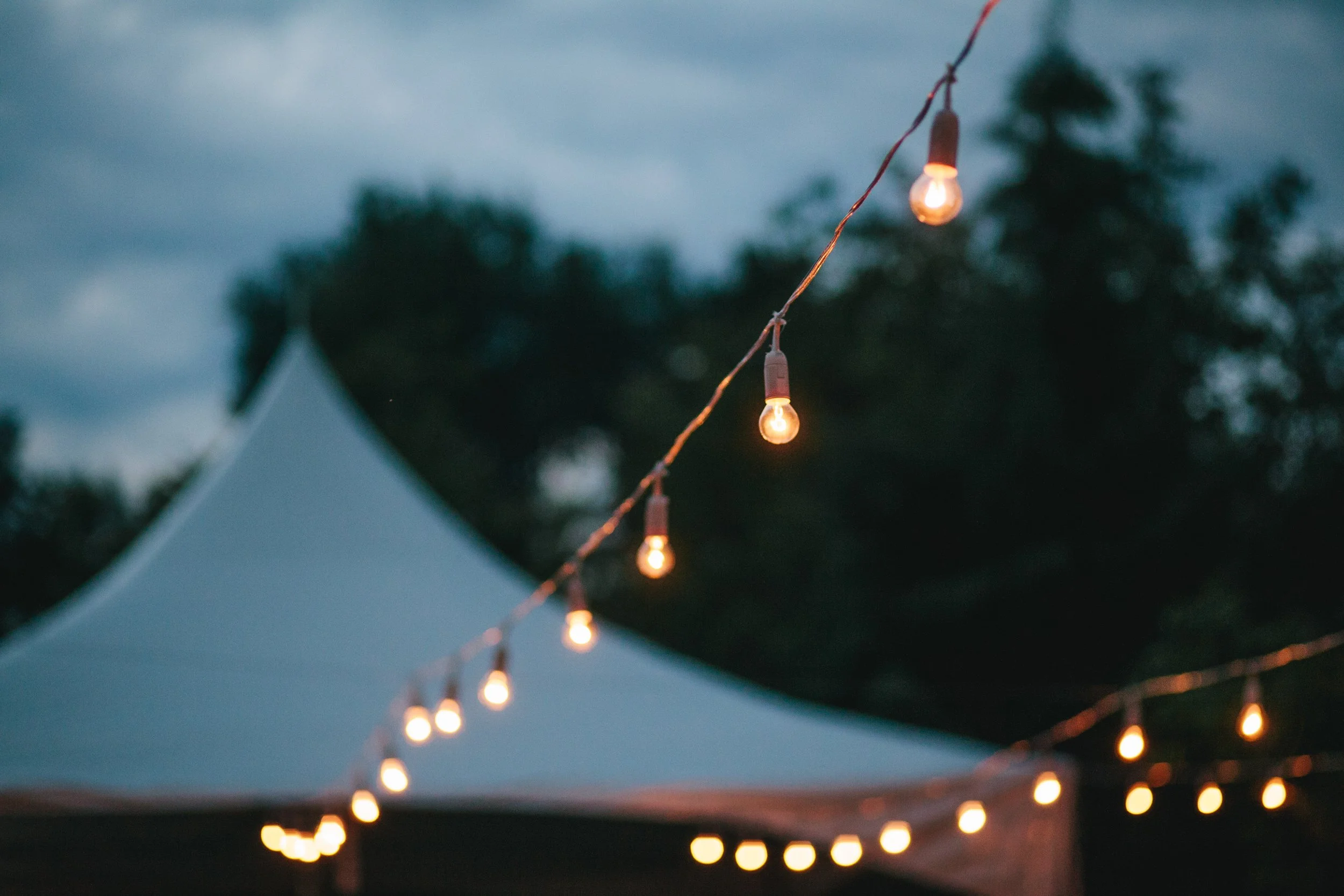 String of outdoor hanging string lights glowing at dusk, with a blurred background of trees and a tent.