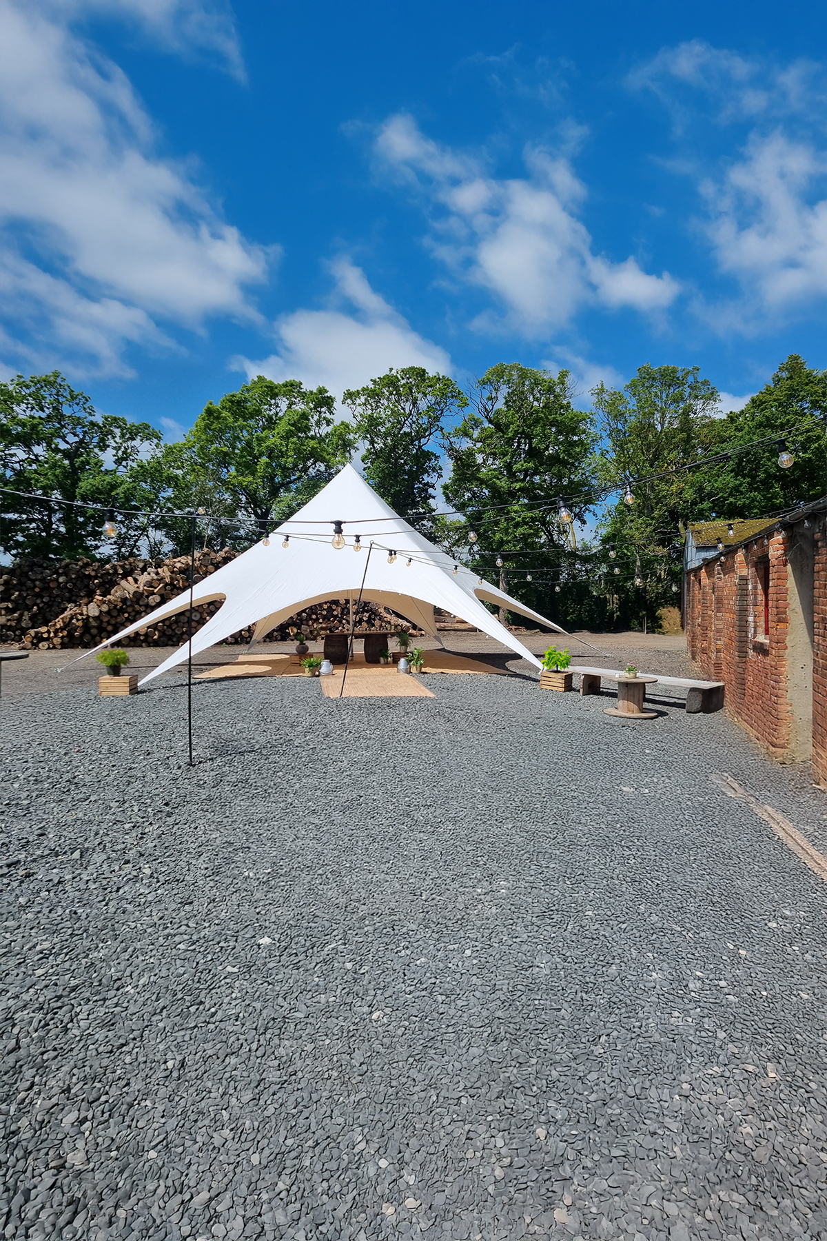 Outdoor event space with a large white tent on gravel ground, surrounded by string lights, potted plants, and rustic benches, with trees and blue sky in the background.