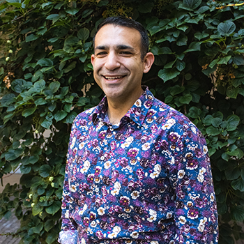 A smiling man wearing a colorful floral shirt standing in front of green leafy plants.