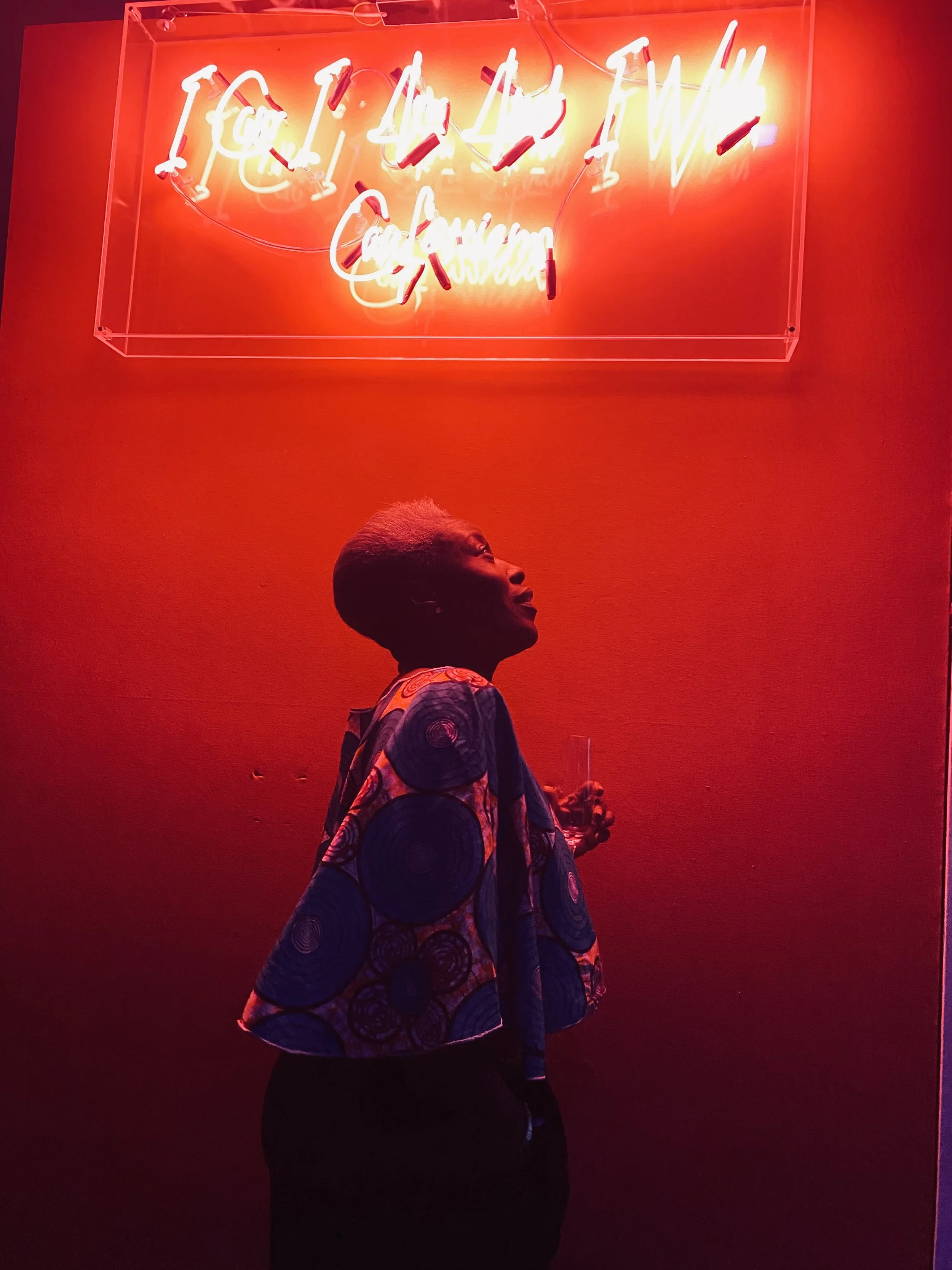 A person standing in front of a red background with a neon sign that reads 'I feel like I do' in cursive. The person is looking up at the sign, wearing a colorful patterned jacket.