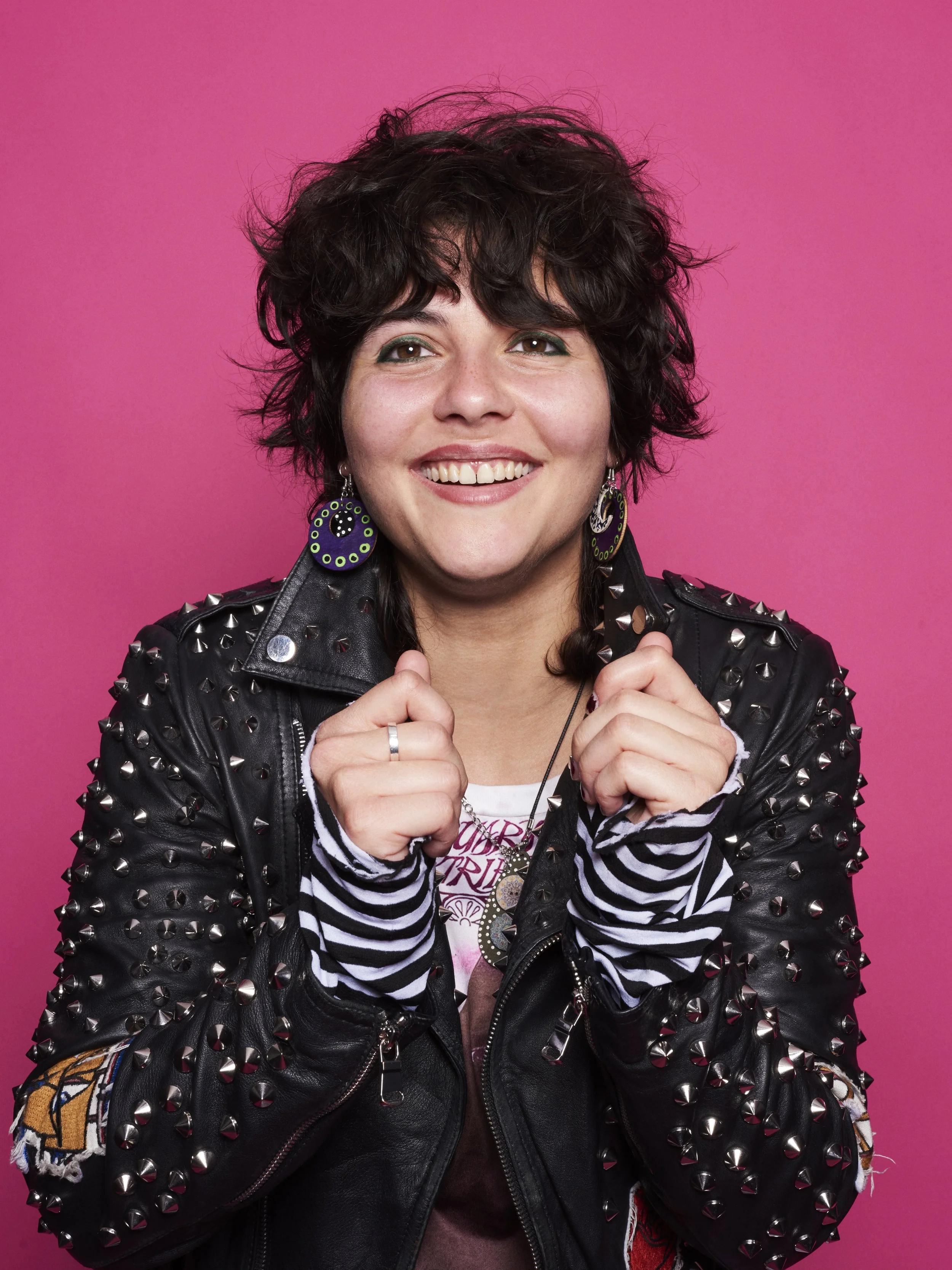A woman with curly dark hair smiling, wearing a studded black leather jacket, striped long-sleeve shirt layered underneath, colorful earrings, and a graphic t-shirt, against a pink background.