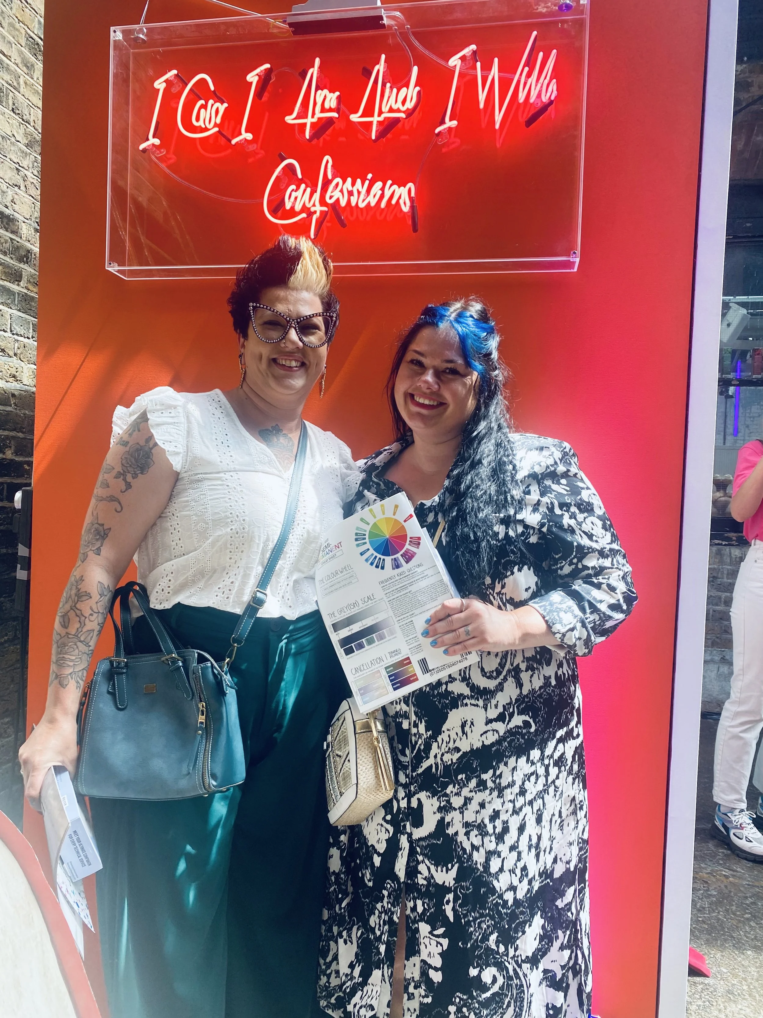 Two women standing together in front of a red wall with a neon sign that reads 'I Care I Am Able I Will' and 'Caregivers.' One woman has colorful hair, glasses, a white top, and tattoos. The other woman has dark hair with blue highlights, a black and