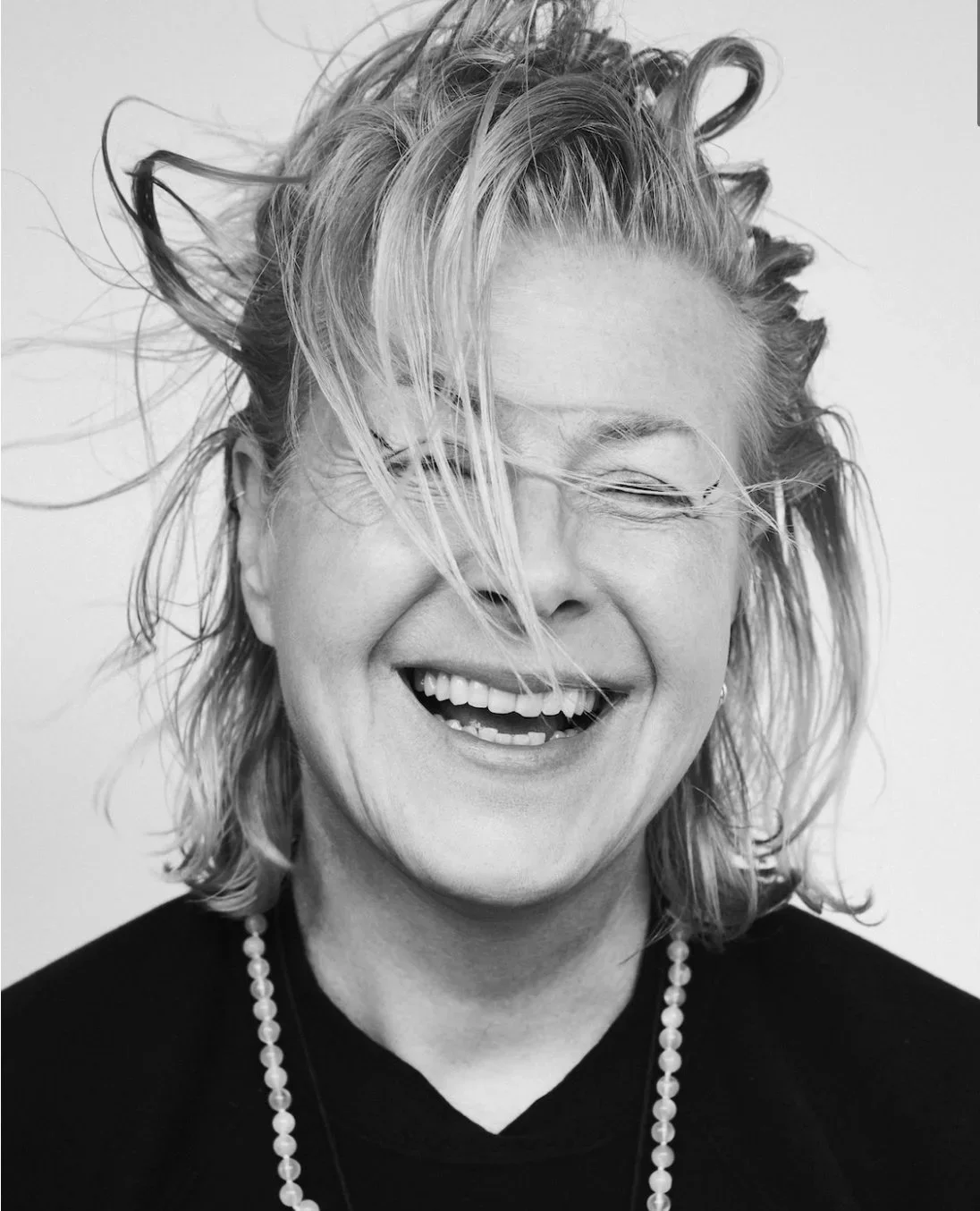 Black and white photo of a woman with messy, shoulder-length hair laughing with her eyes closed, wearing a pearl necklace and a dark top.