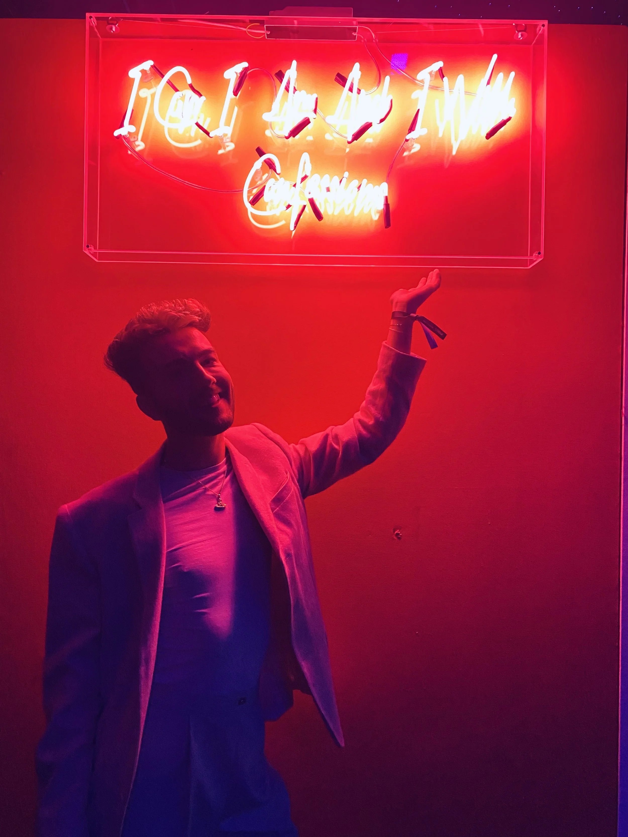 A person smiling and pointing at a bright red neon sign on a red wall. The sign reads 'TOO SEXY TO BE OK' in cursive script.