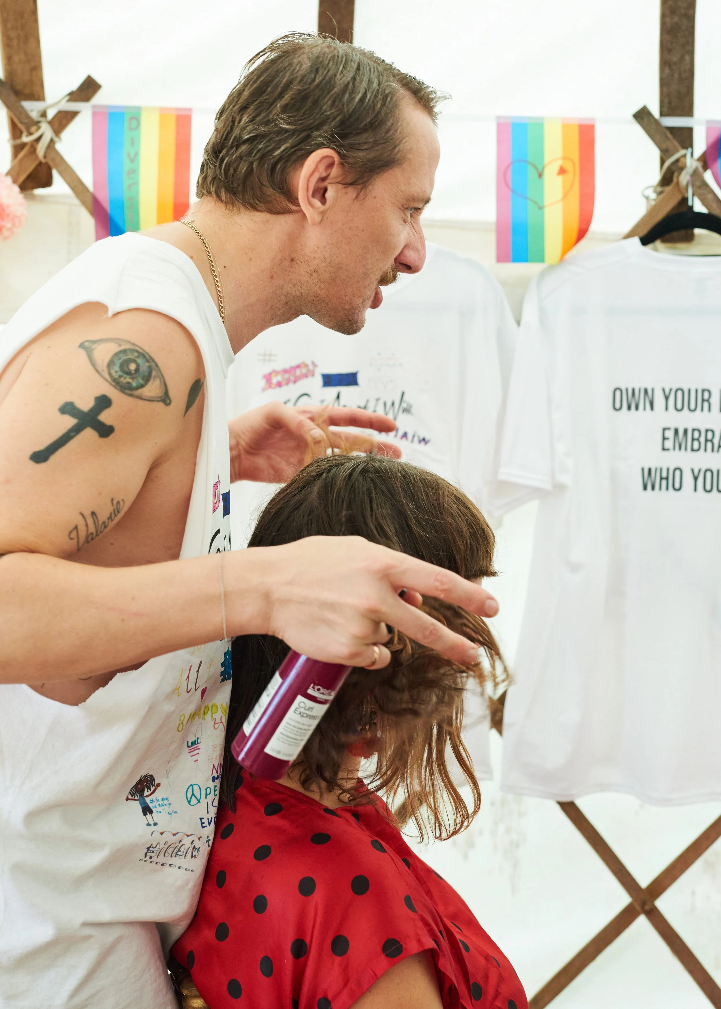 A man with tattoos gives a woman a haircut at a booth with clothing and rainbow pride flags.