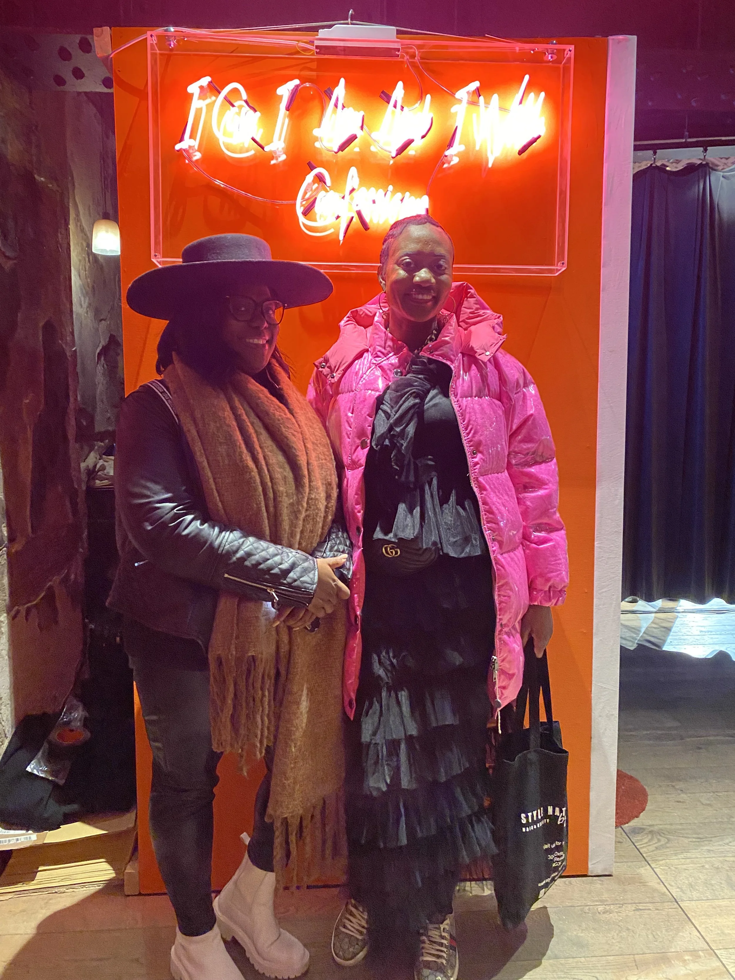 Two women standing in front of an orange wall with a neon sign that says 'ITALIAN.' One woman is wearing a large black hat, glasses, a black leather jacket, and a brown scarf. The other woman is wearing a bright pink puffy jacket and a black dress wi