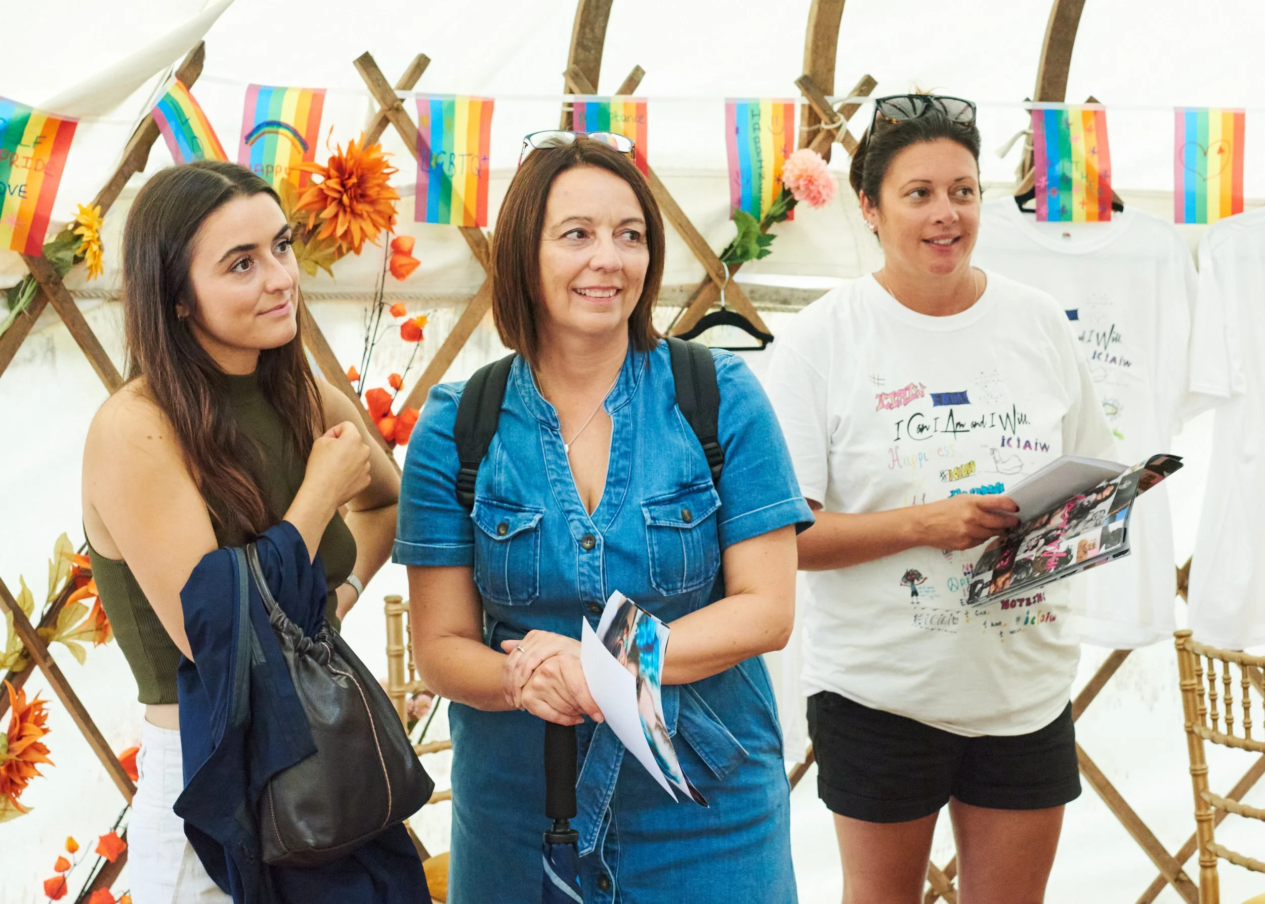 Three women standing inside a decorated event tent, looking at something off-camera. The background has vibrant rainbow-themed decorations, flowers, and T-shirts on display.