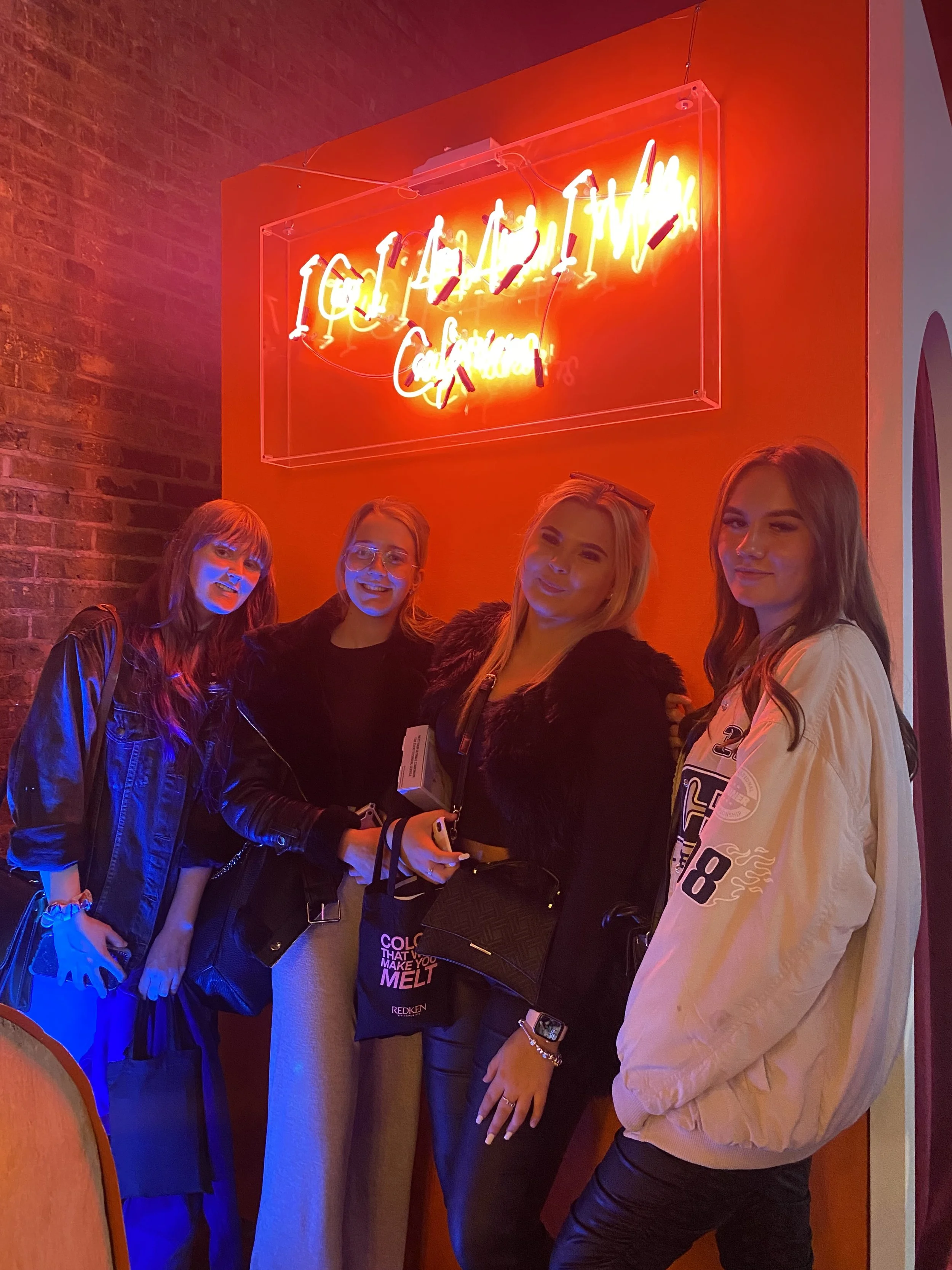 Four women standing in front of a neon sign that reads 'CHADWICK COUTURE' inside a restaurant or lounge. They are dressed casually and smiling.