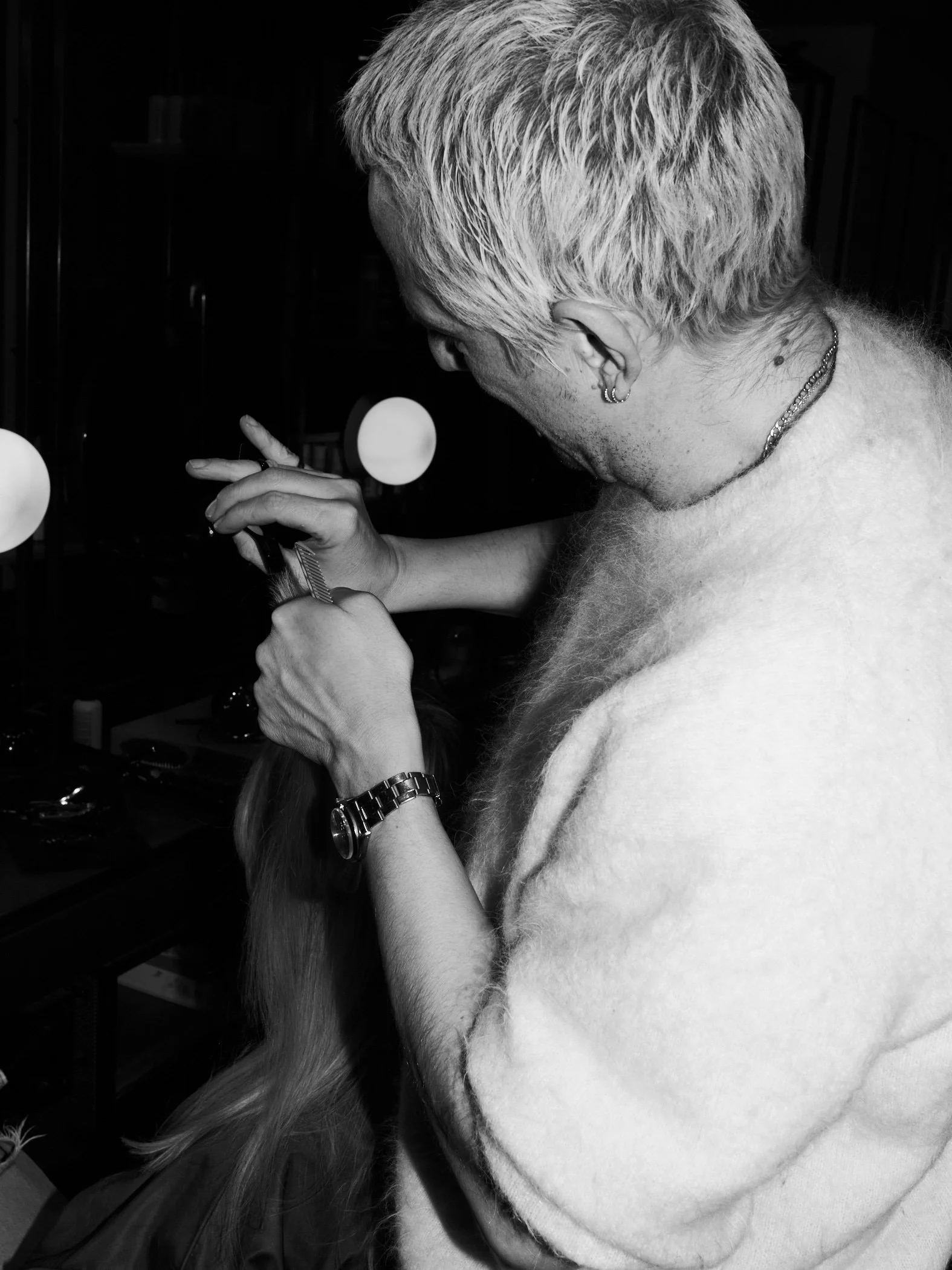 A person with short, light-colored hair and multiple piercings styles another person's long hair using a comb in a salon or grooming setting.