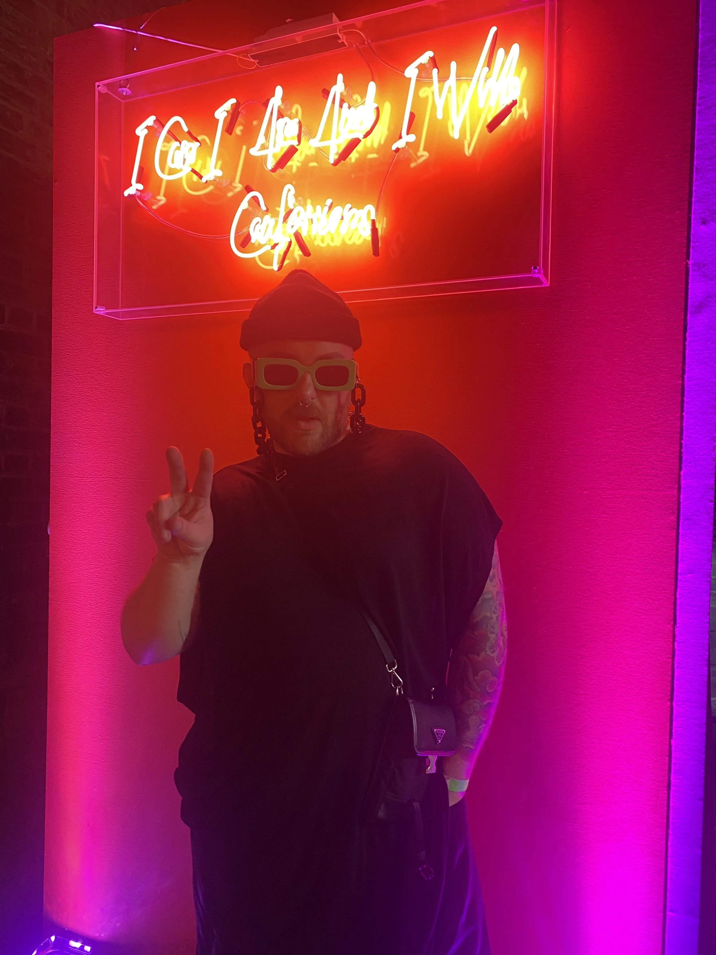 Person standing in front of a neon sign with bright pink and orange lights, wearing large sunglasses, a beanie, and black clothing, making a peace sign gesture.