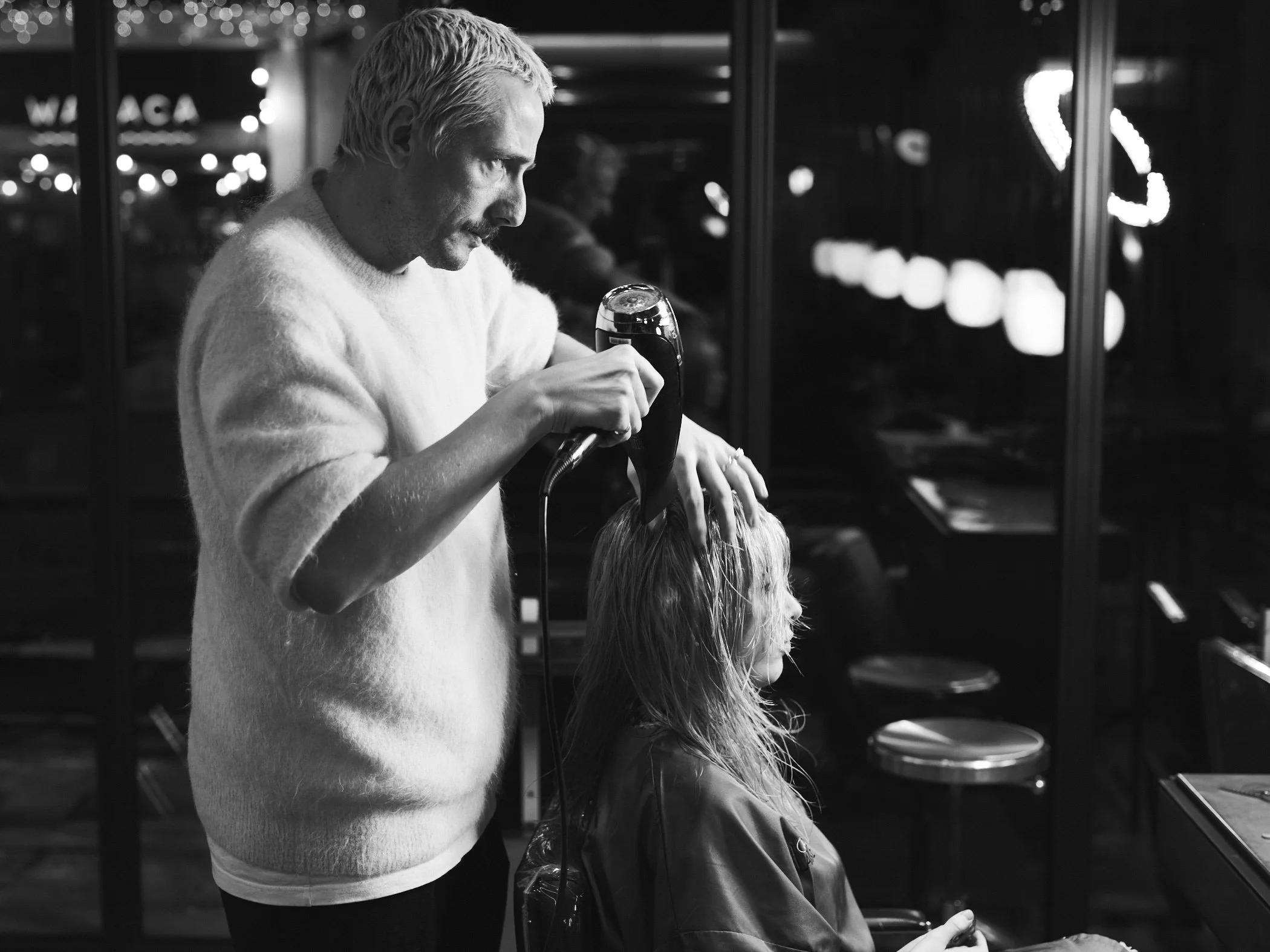 A man is drying a woman's hair inside a dimly lit salon at night, with reflections in the windows.