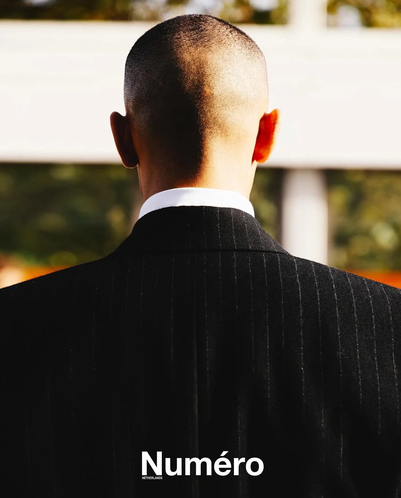 Back of a man in a black pinstripe suit, facing away, with a shaved head and a white shirt collar visible