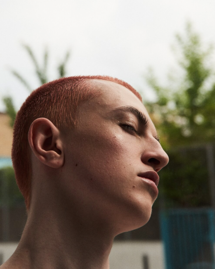 Close-up of a shirtless young person with short red hair, wearing a shirtless expression, outdoors with trees and a fence in the background.