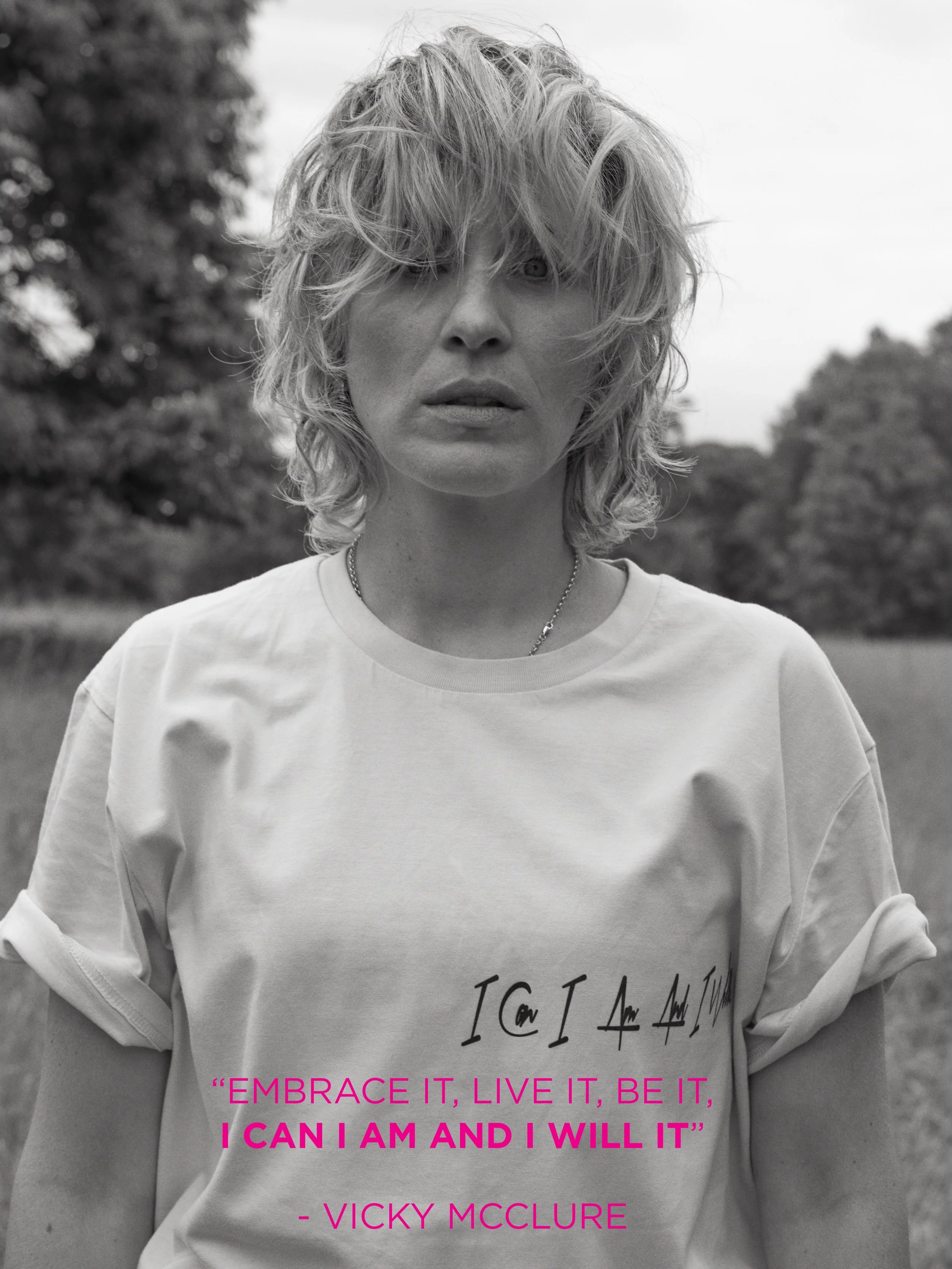 A black and white photo of a woman with wavy, disheveled hair, wearing a T-shirt with a motivational quote and standing outdoors with trees in the background.