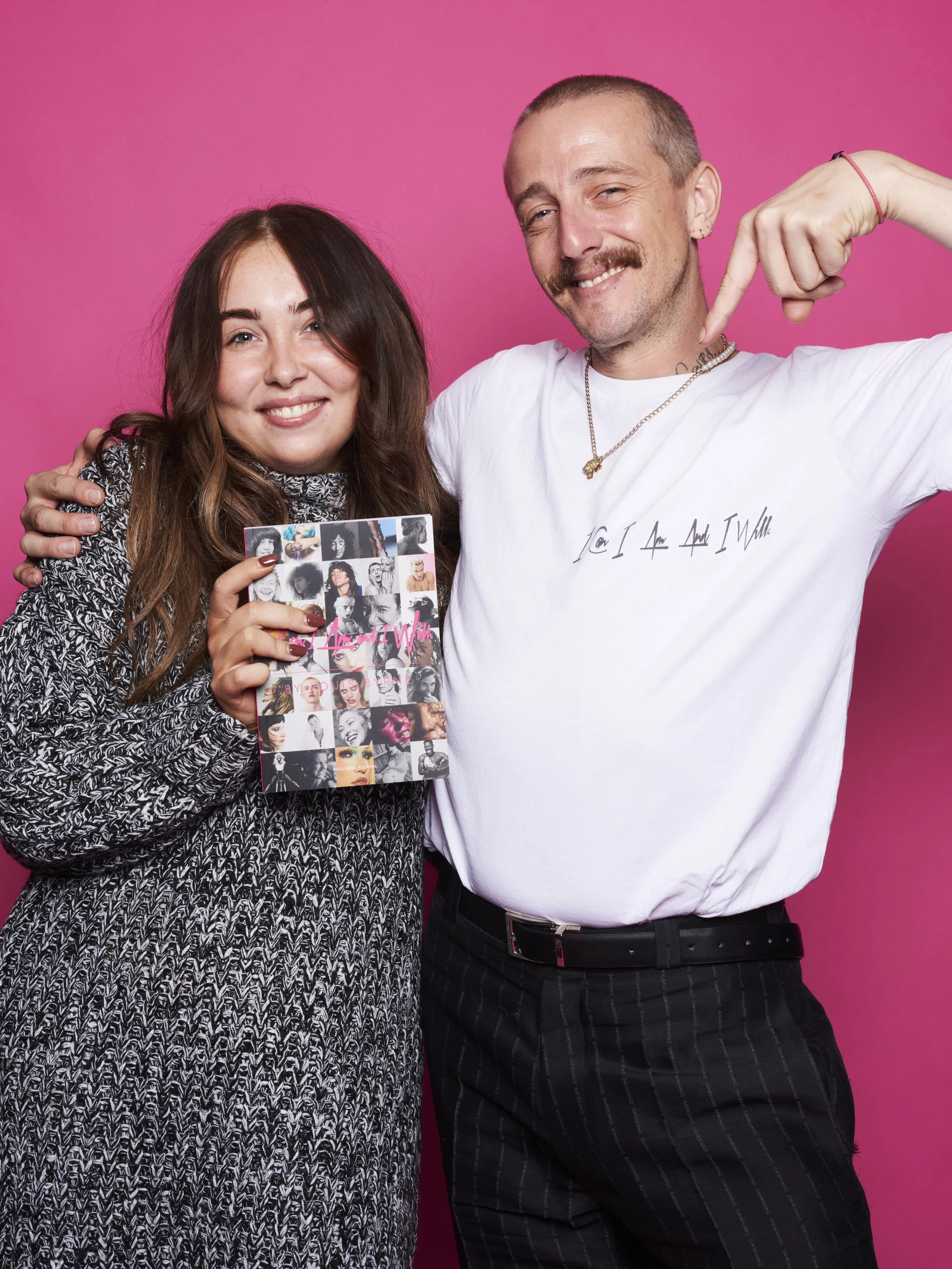 A woman and a man smiling and posing for a photo against a pink background. The woman is holding a book with a collage of black and white and color photos of women. The man is pointing to the woman and wearing a white t-shirt with hand-written style