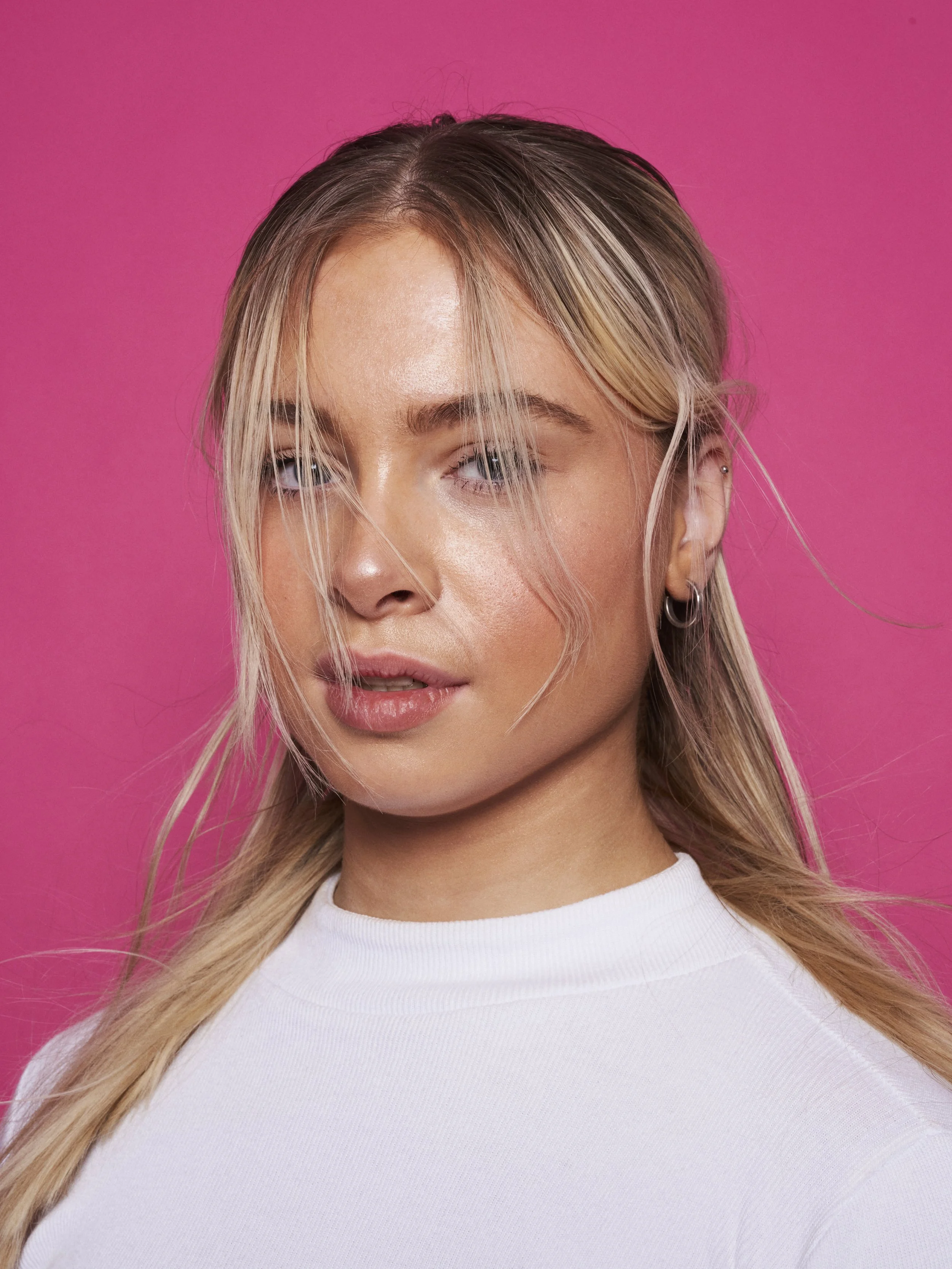 Close-up portrait of a young woman with blonde hair, blue eyes, wearing a white top, posed against a pink background.
