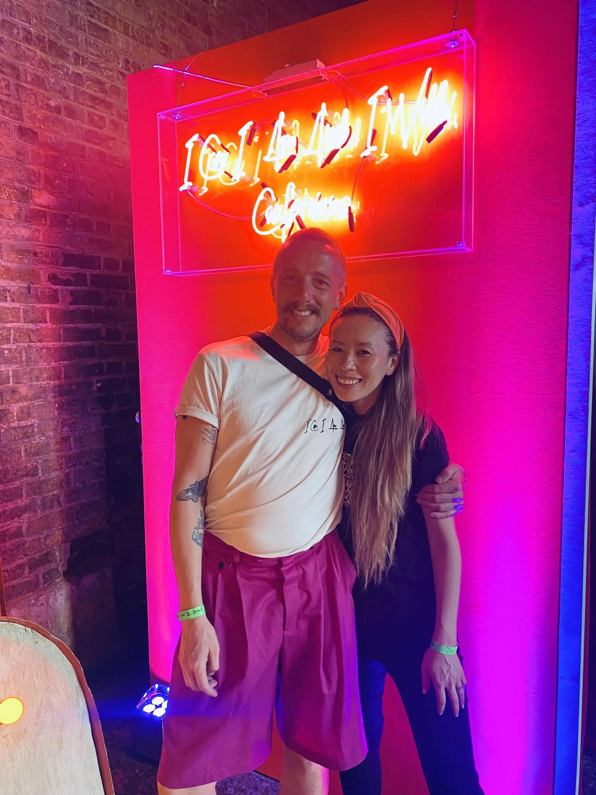 Two smiling people, a man and a woman, posing in front of a neon sign that reads 'ICU and MIAMI Culture' at a nightclub or bar, illuminated with pink and purple lighting.