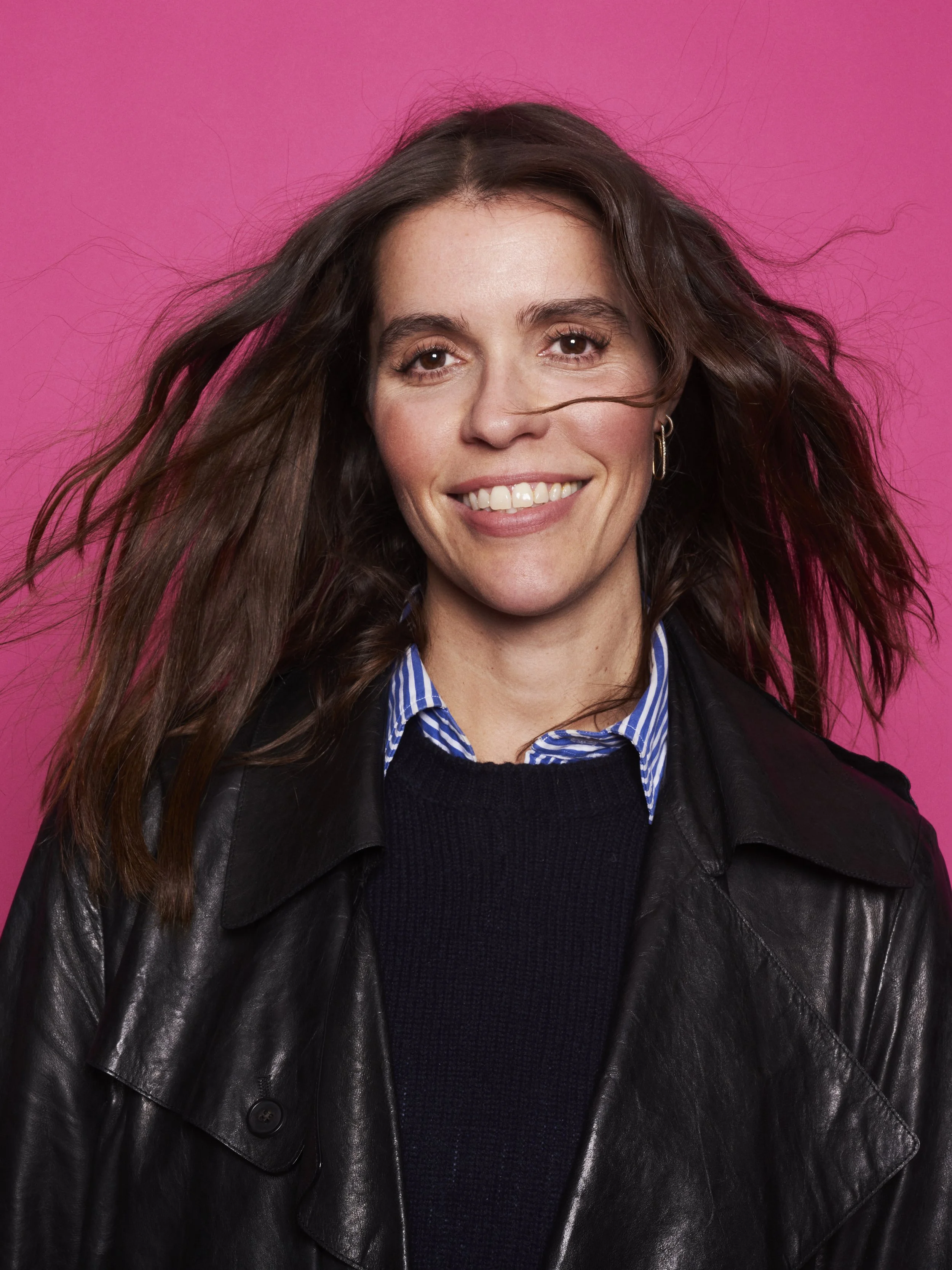 A smiling woman with shoulder-length brown hair, wearing a black leather jacket over a blue and white striped shirt, against a pink background.