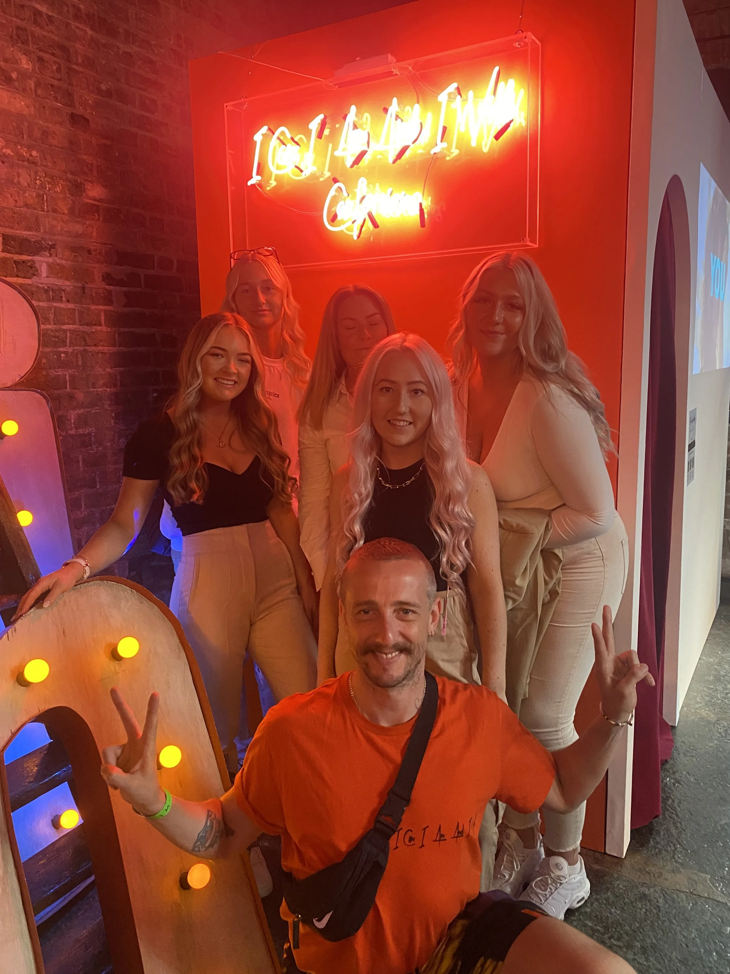 A group of six friends, five women and one man, posing for a photo in front of a neon sign that reads 'I GOT A TICKET'. The man is sitting in front, smiling and making peace signs with both hands. The women are standing behind him, smiling and lookin