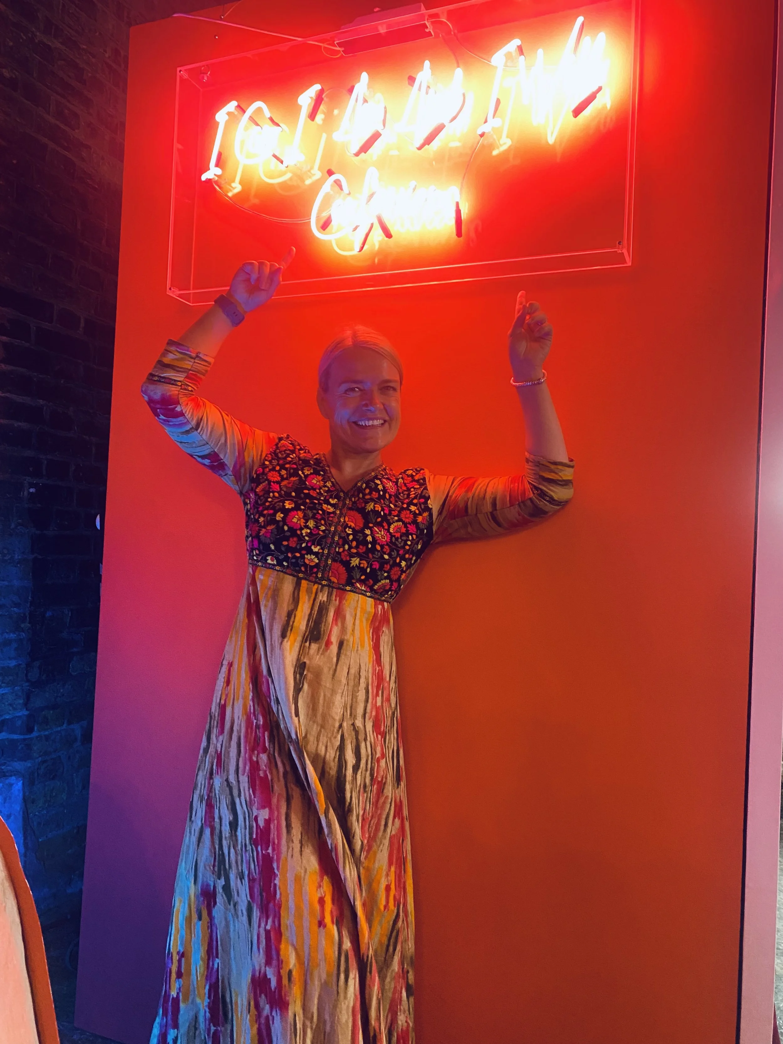 A woman standing in front of a red wall with a neon sign that reads 'Ice Cream' in cursive. She is smiling and holding her arms up in a celebratory pose.