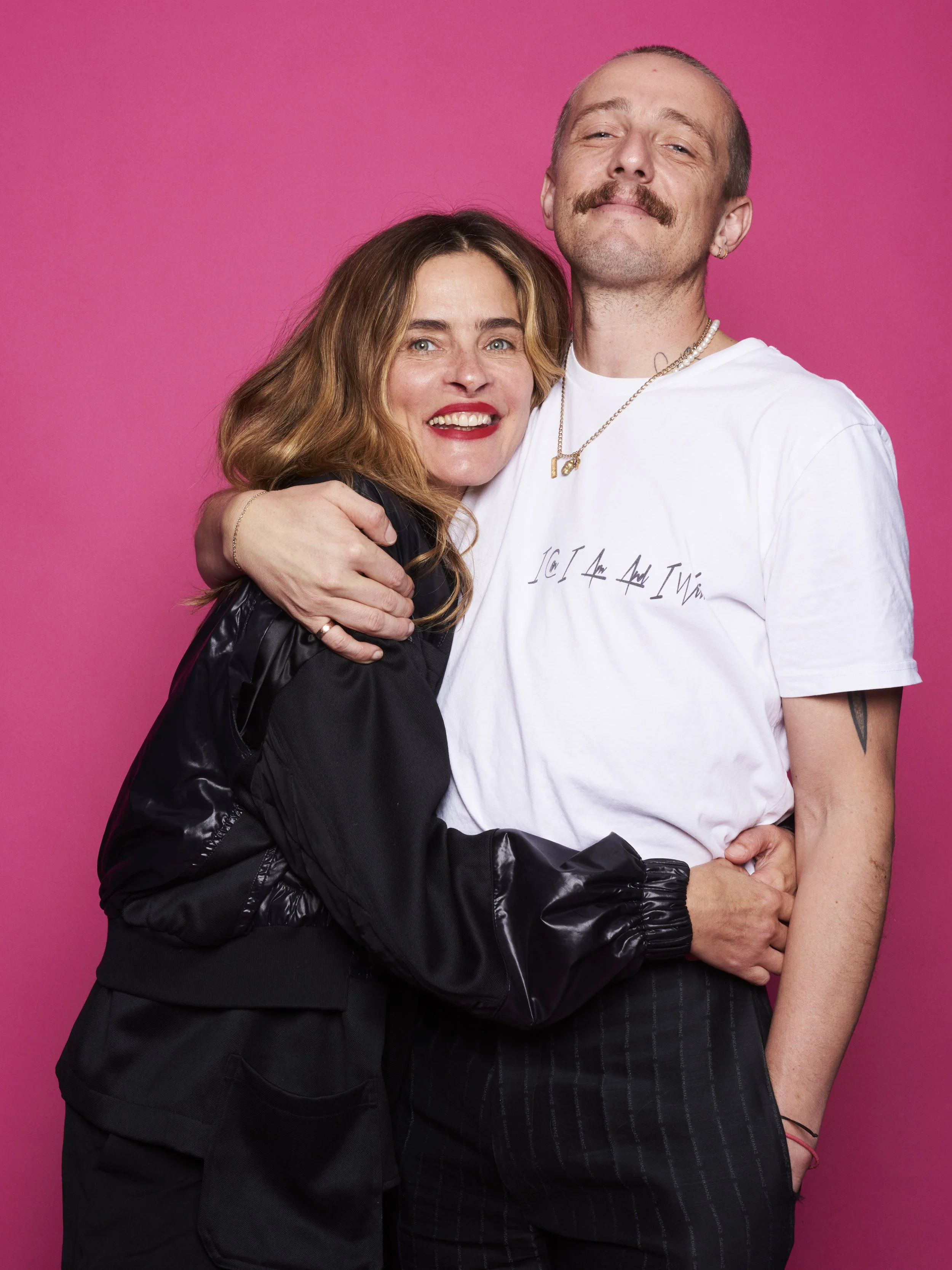 A woman and a man hugging against a pink background. The woman has light brown, wavy hair and is wearing a black jacket. The man has a mustache, is bald, and is wearing a white t-shirt with text, earrings, a necklace, and striped pants.