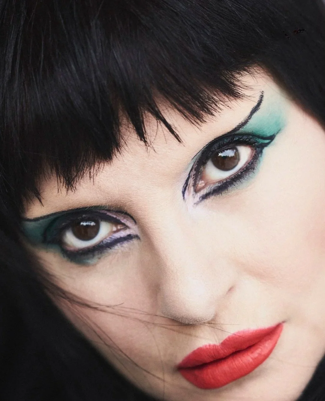 A close-up of a woman's face with bold, artistic makeup, including turquoise and black eye makeup and red lipstick.