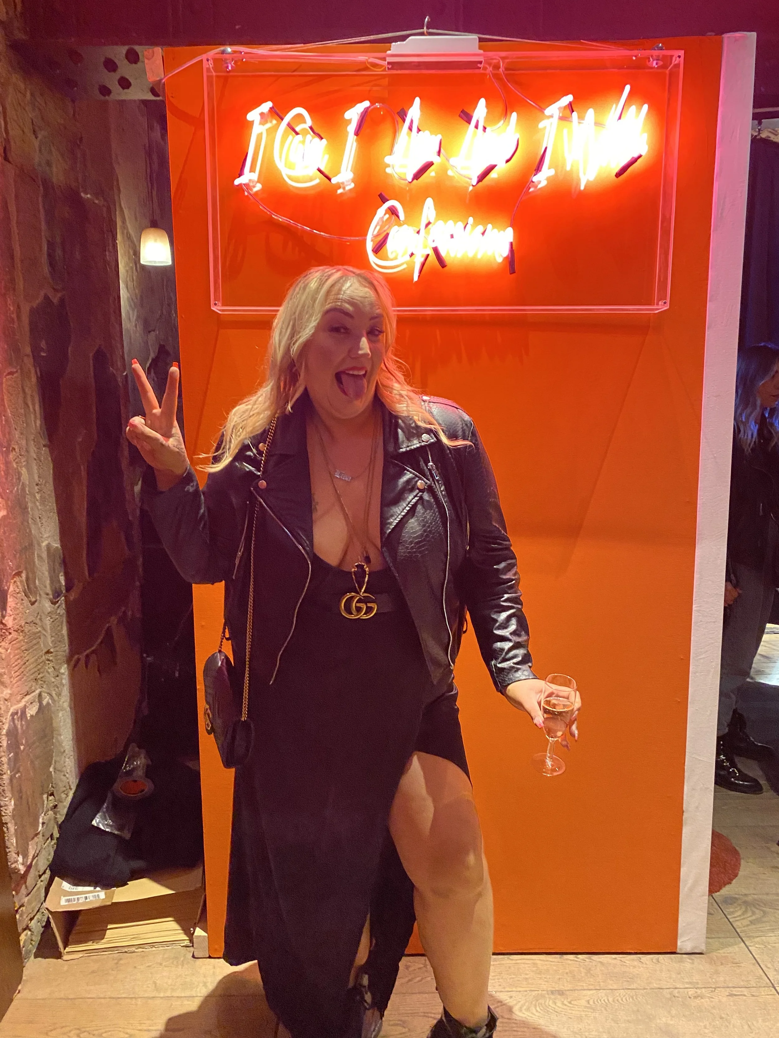 A woman with blonde hair wearing a black leather jacket, a black dress with a high slit, and multiple necklaces, posing with a peace sign and holding a glass of white wine in front of a bright orange wall with neon sign that says 'If I Had A Kitchen'