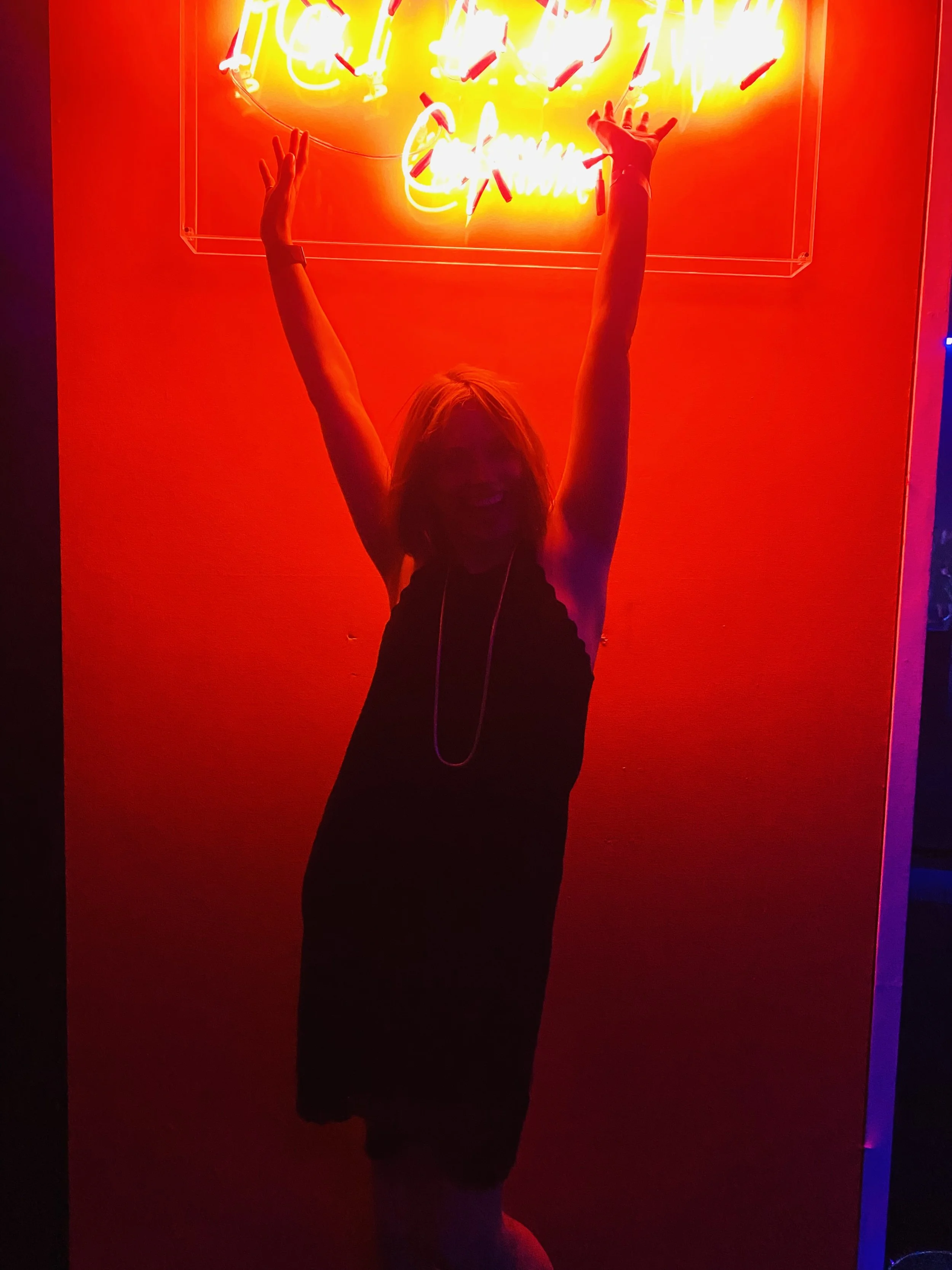 Person with arms raised in front of a bright neon sign that reads 'Hello It's Nice to See You' in yellow and white, against a red background.