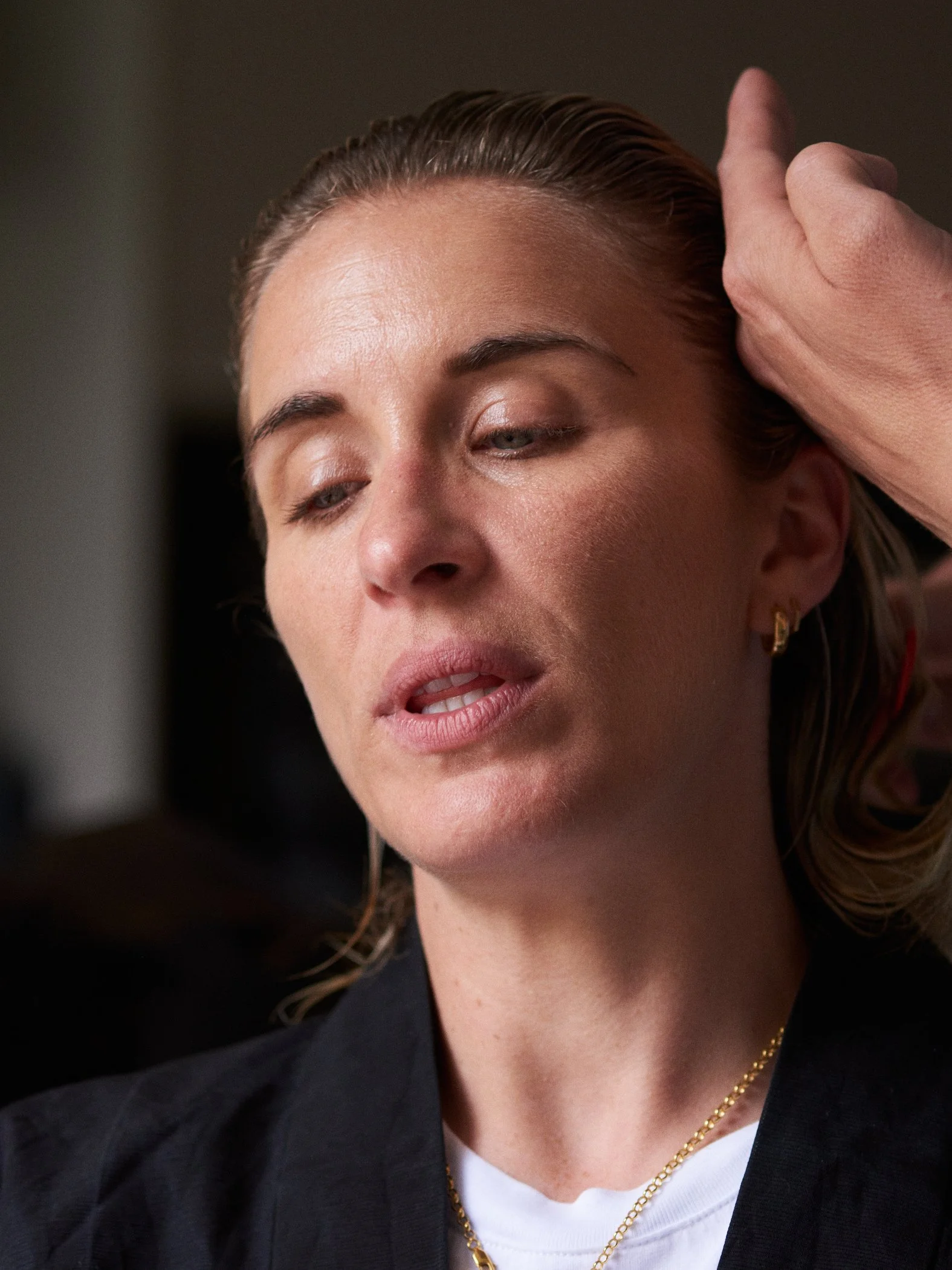 A woman with slicked-back hair and gold earrings has her head touched by a hand, appearing to receive a massage or therapy, in a close-up shot.
