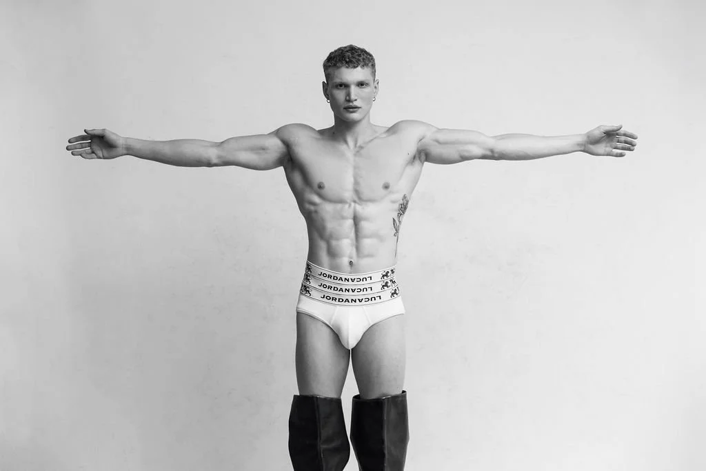 A shirtless young man standing with arms outstretched against a plain background, wearing white underwear with "JORDAN AVEN" written on the waistband and black thigh-high boots.