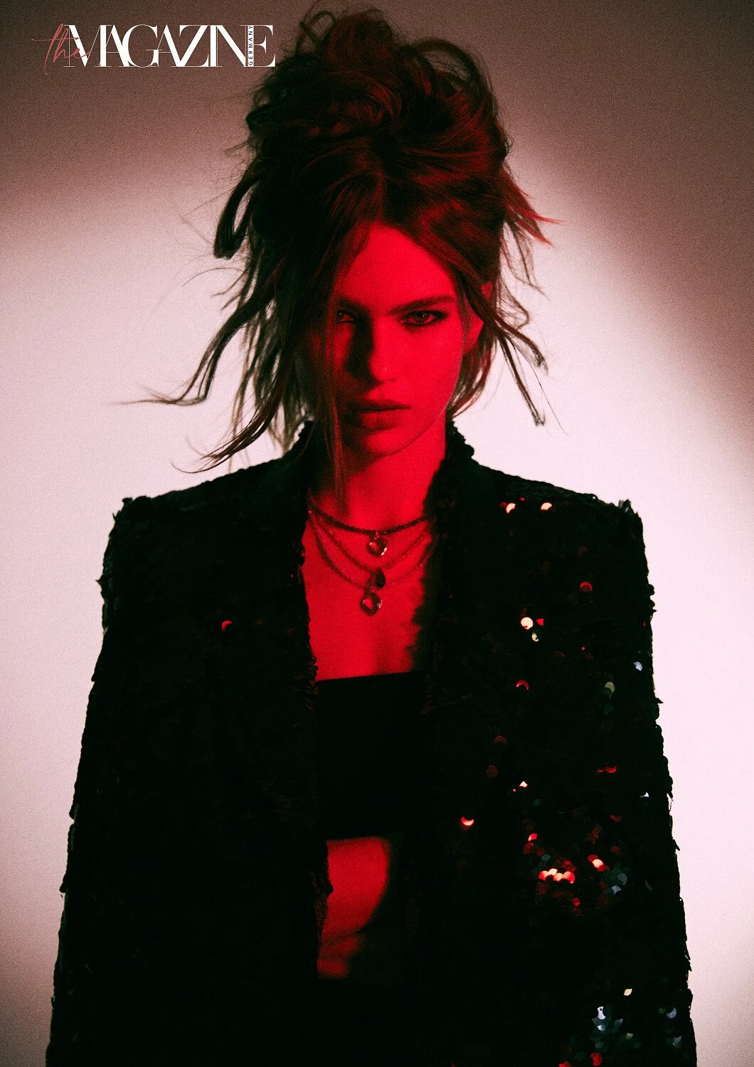 A woman with voluminous, messy hair wearing a black sequined jacket and layered necklaces, posing against a gradient background with a red light cast on her face.