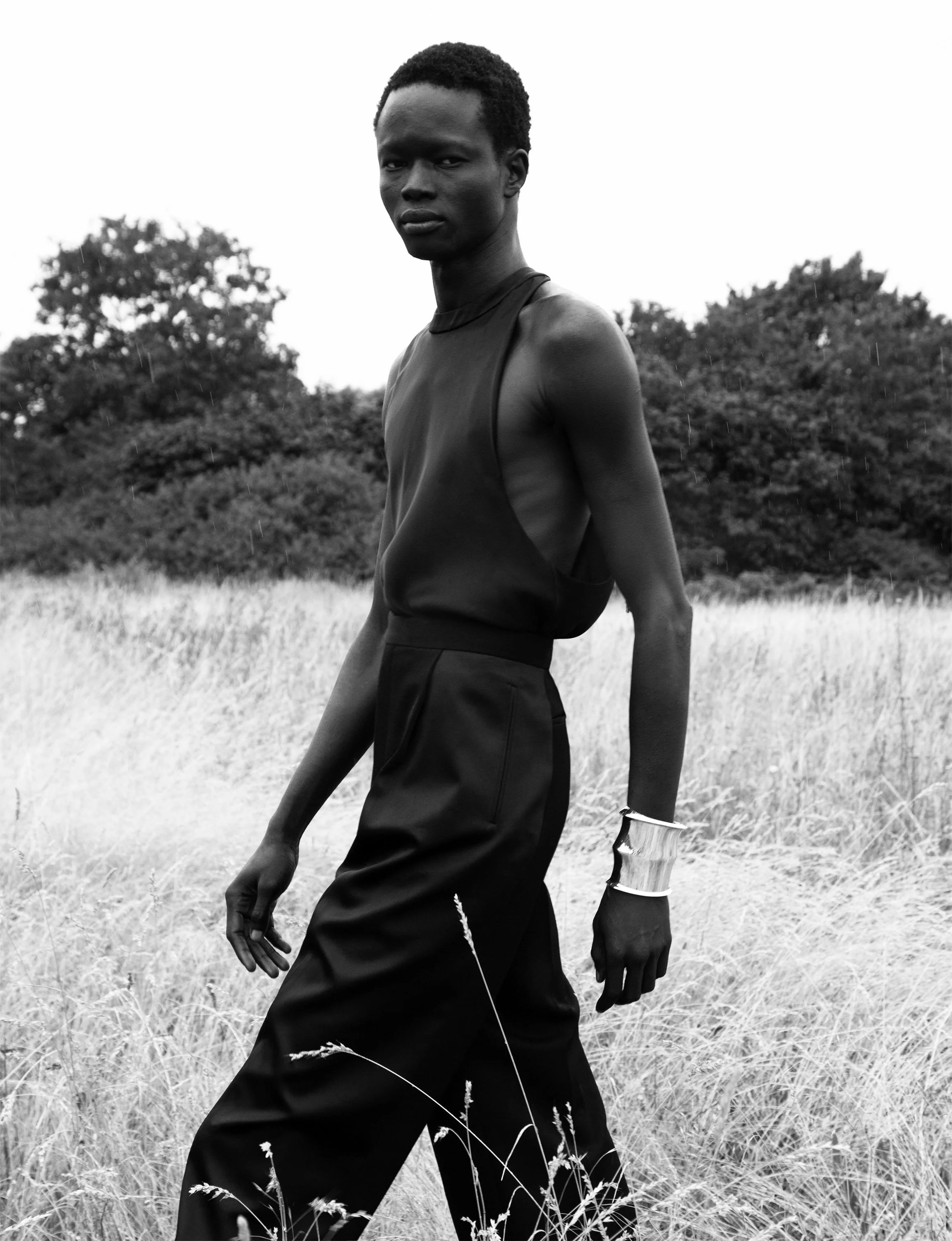 Black and white photo of a slender person walking through a field with trees in the background, wearing a sleeveless top, wide-leg pants, and a bracelet.