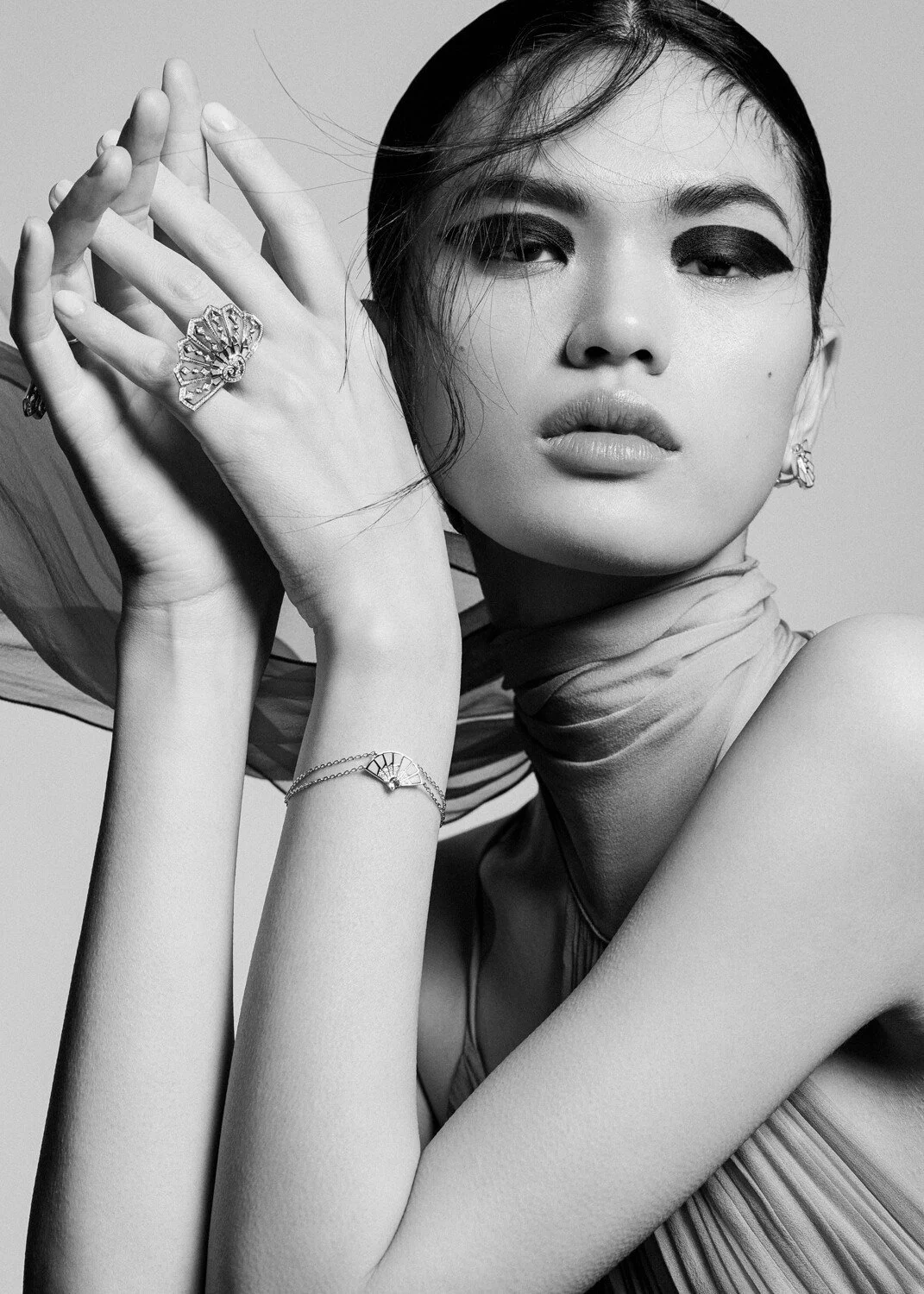 Black and white photo of a woman modeling jewelry, including rings, earrings, and a bracelet, with her hands near her face and her hair styled with a headscarf.