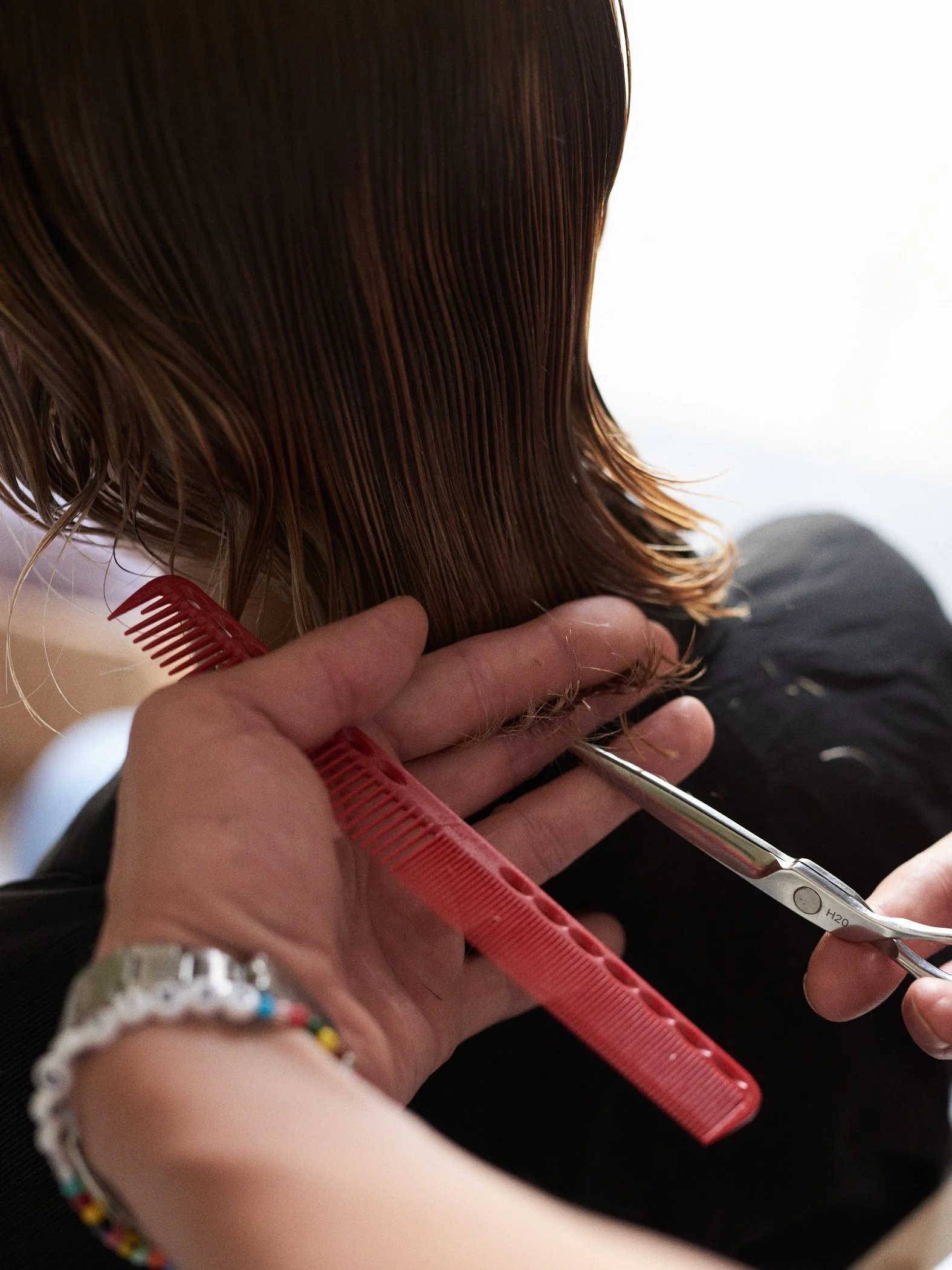 A person getting a haircut with scissors and a red comb, hairstyle with brown wet hair, in a salon setting.