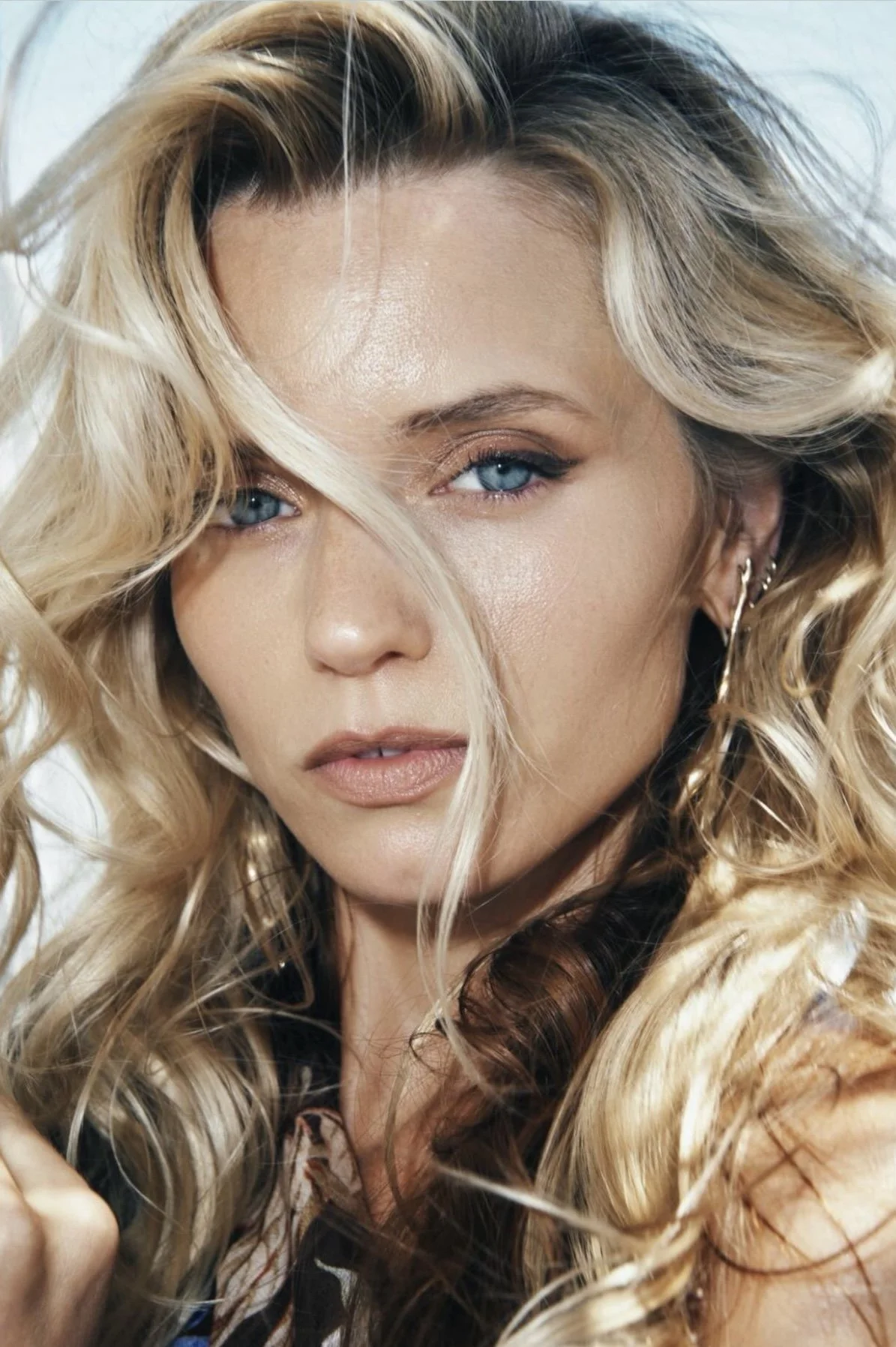 A close-up of a woman with wavy blonde hair, blue eyes, and makeup, looking directly at the camera.
